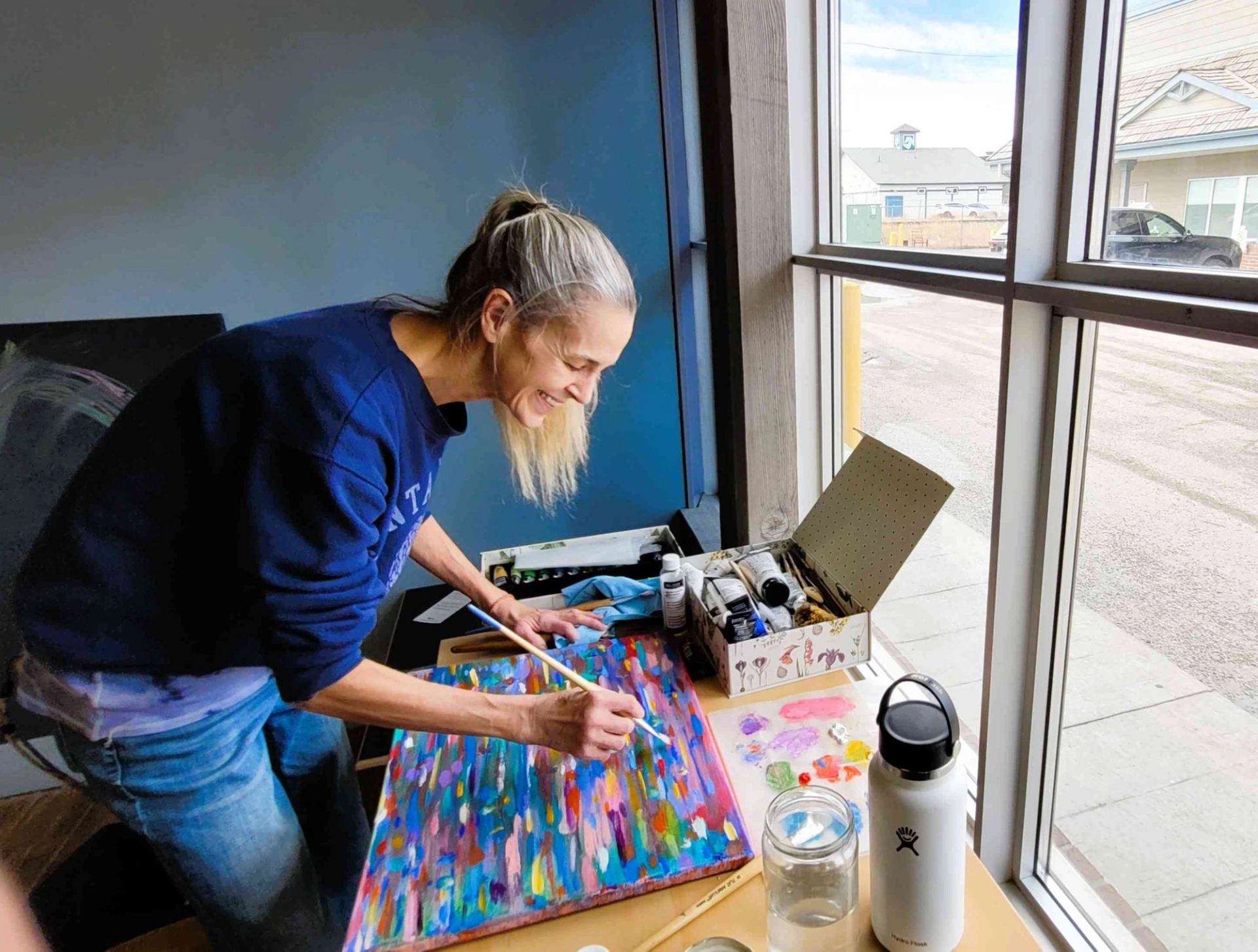 Artist painting a colourful abstract canvas beside a large window with art supplies.