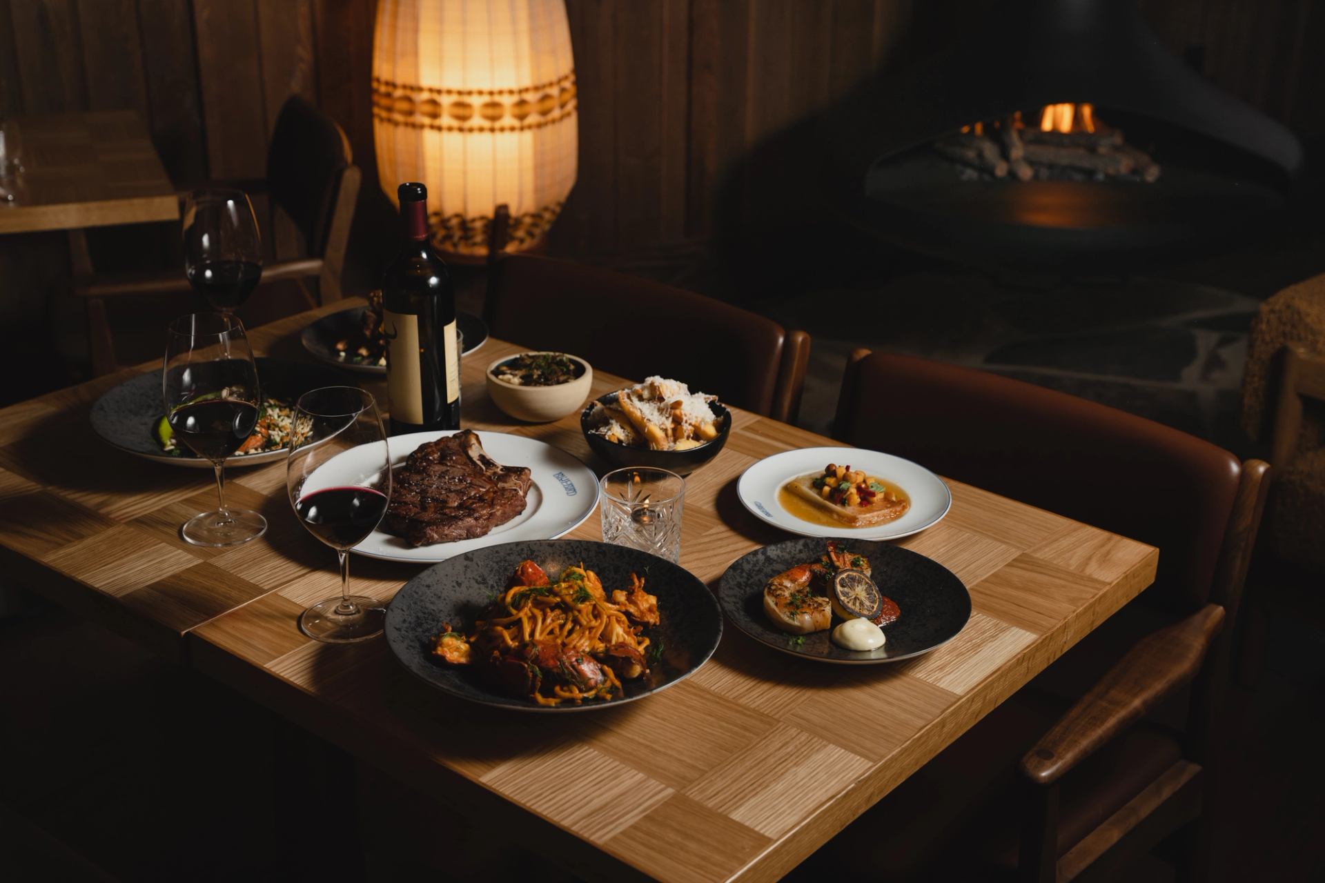 A table set with steak, pasta, shared dishes, and wine in a warm restaurant setting.