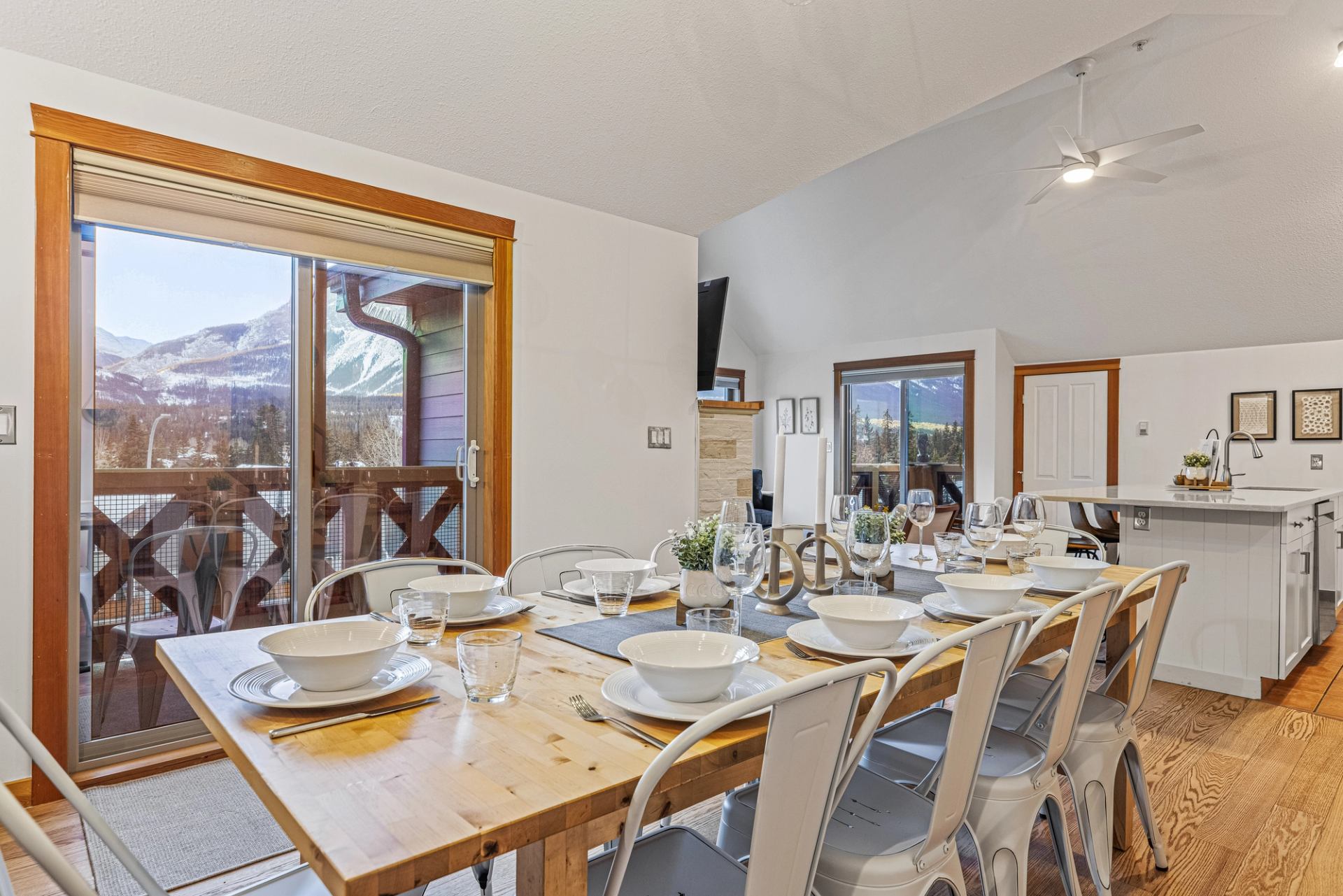 Dining table set with white dishes in open kitchen, with mountain view through windows.