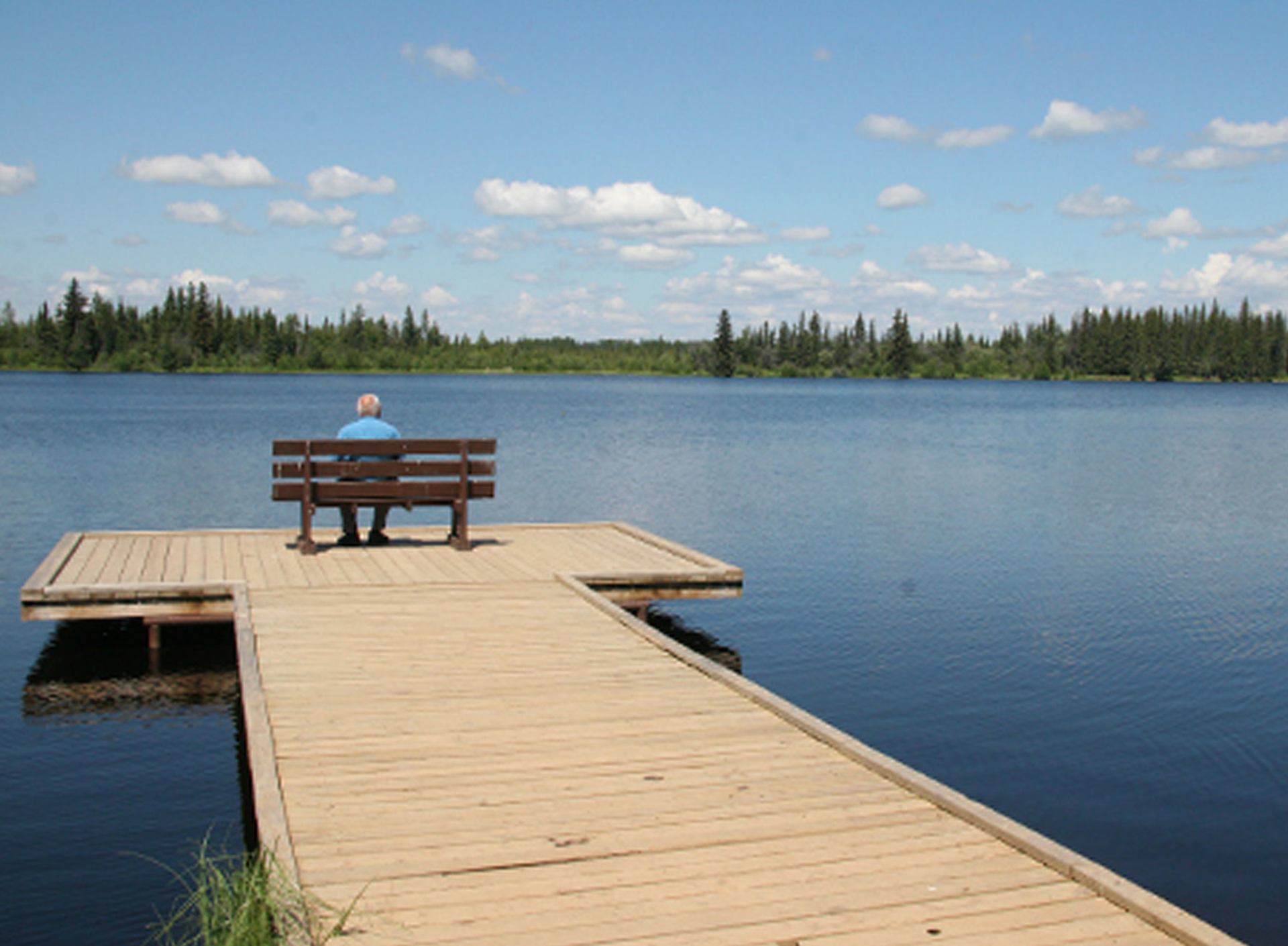 Person on bench at dock overlooking calm lake and forested shore.