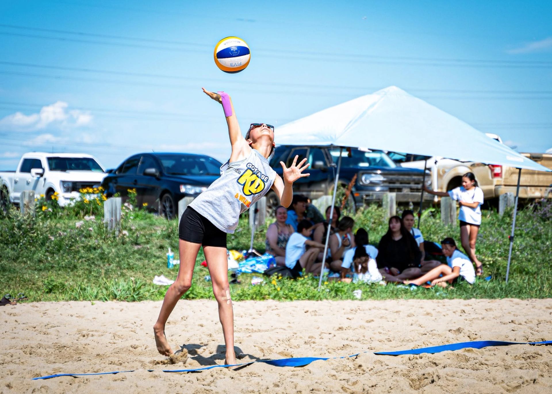 Beach volleyball player jumps to serve ball as spectators sit under canopy nearby.