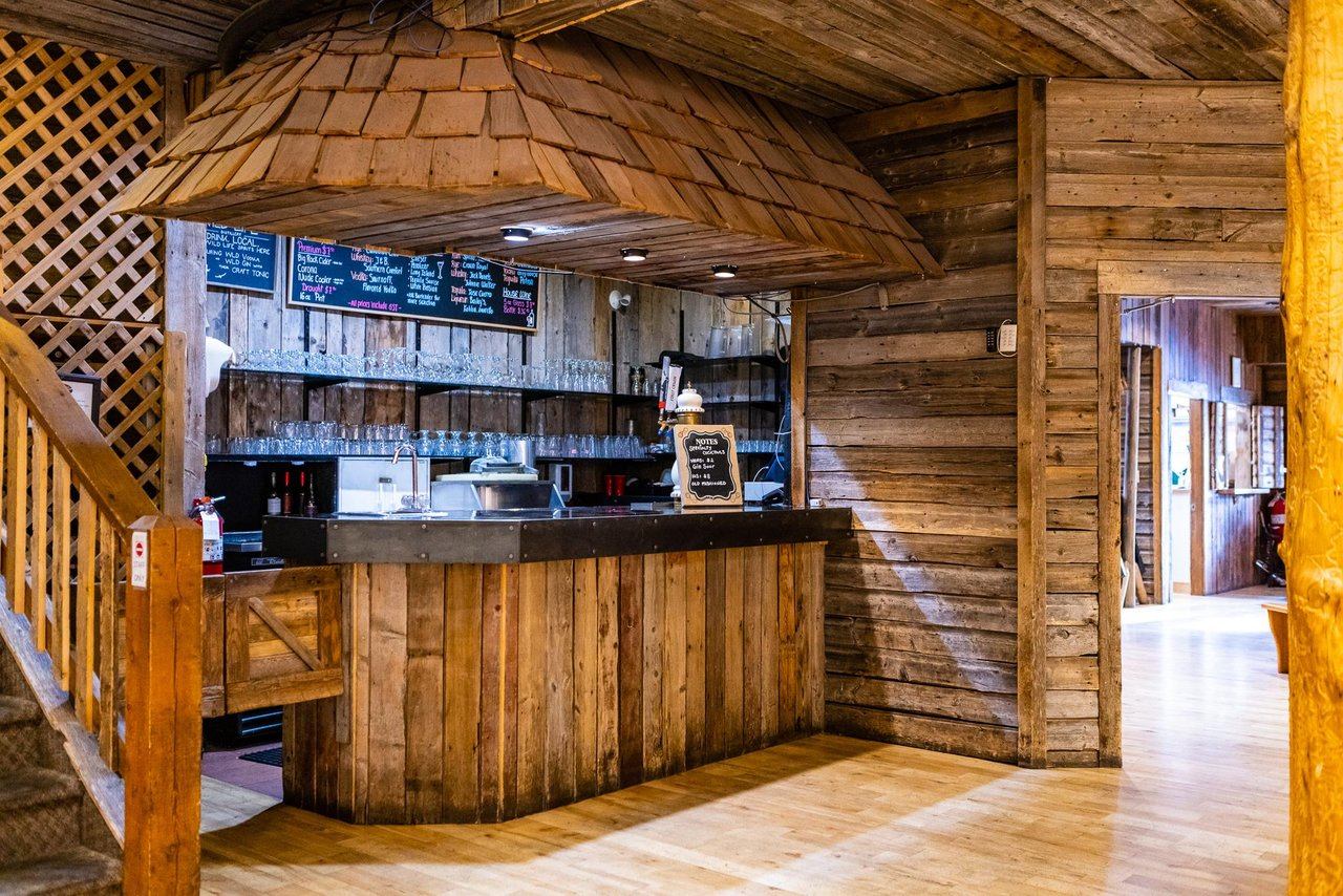 Wooden bar with glassware and a chalkboard menu inside a rustic theatre space.
