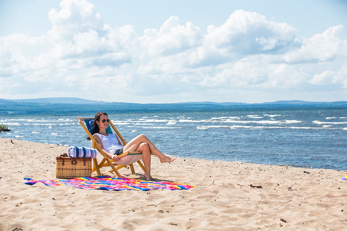 Woman sunbathes on sandy beach with Lesser Slave Lake and Pelican Hills in the background, enjoying a peaceful moment.
