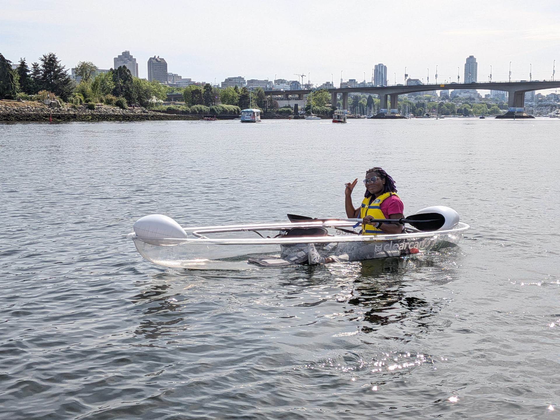 Person in clear kayak on urban waterway with bridge and city skyline.