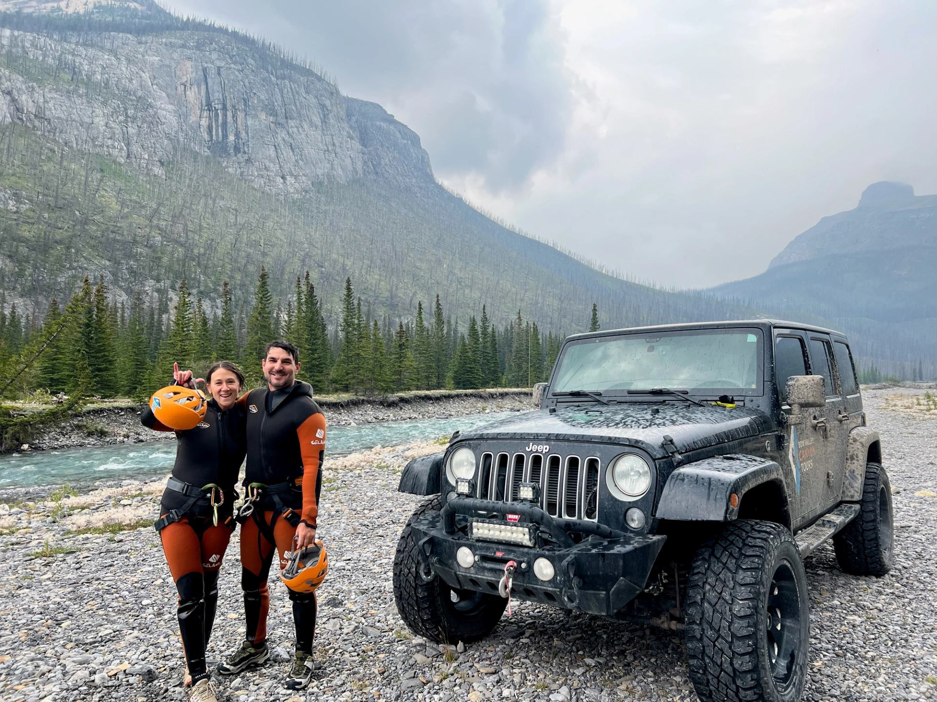 Two adventurers in canyoning gear standing beside an off-road Jeep near a river and mountains.