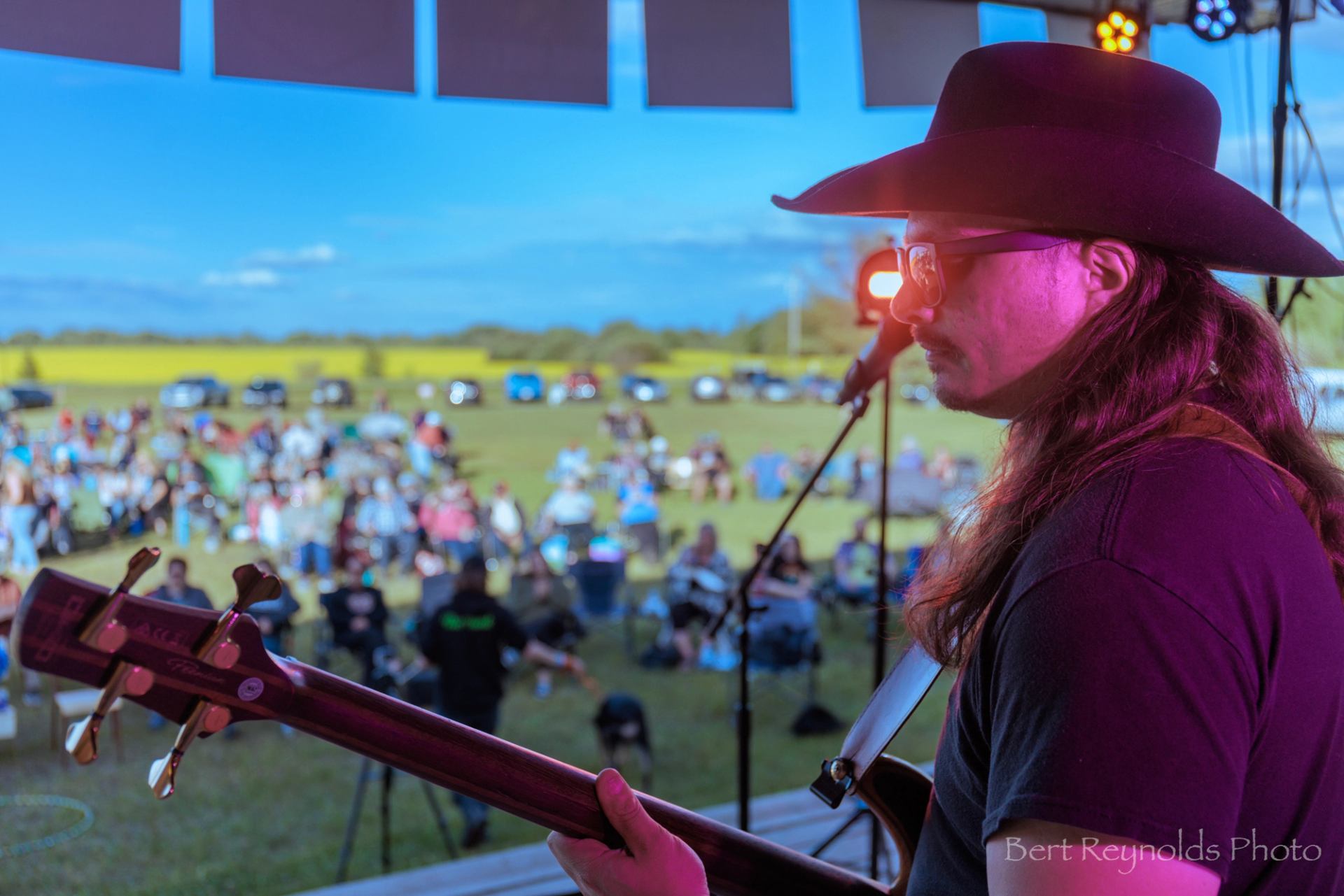 Guitarist in cowboy hat on outdoor stage, audience seated on grass in background.