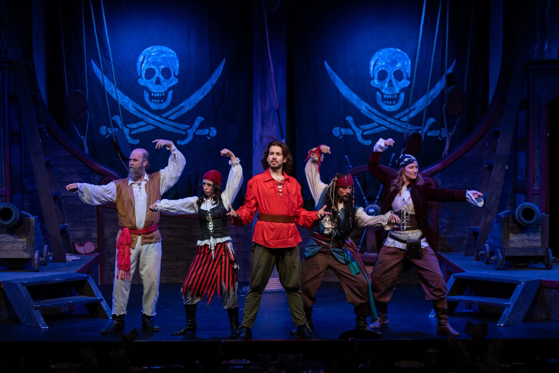 Stage actors in pirate costumes pose under blue skull-and-crossbones backdrops on ship set.