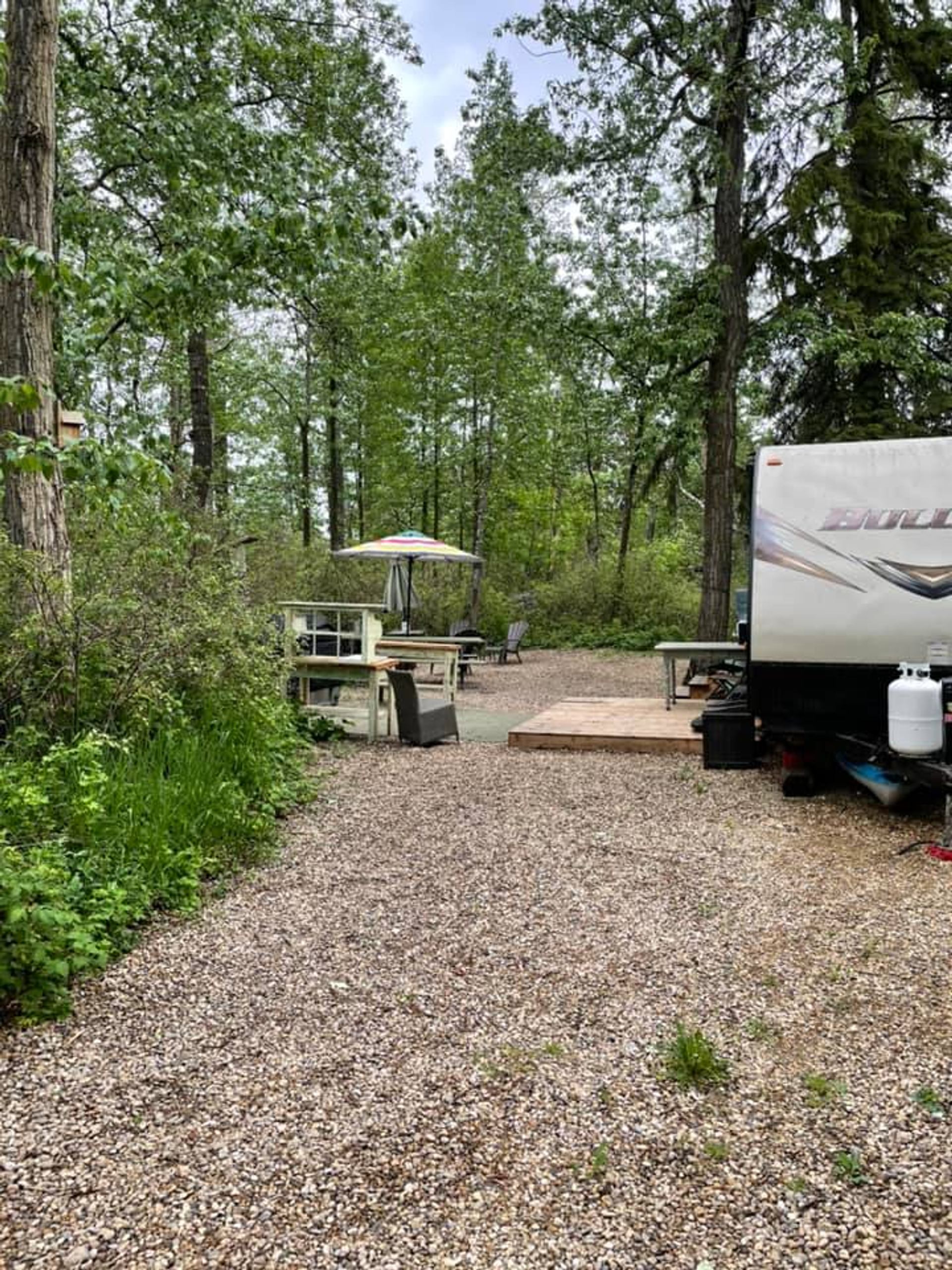 A camper trailer is parked at a wooded campsite with deck seating at Wilson’s Beach Campground.