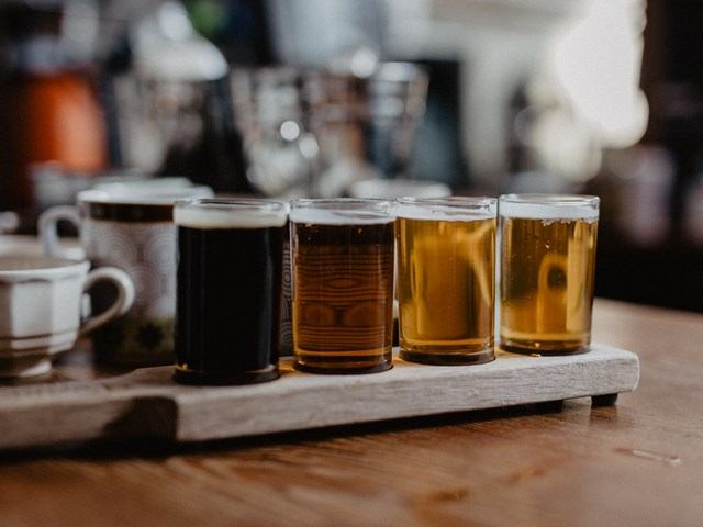 Beer flights in glasses on a wooden table, with mugs in the blurred background.