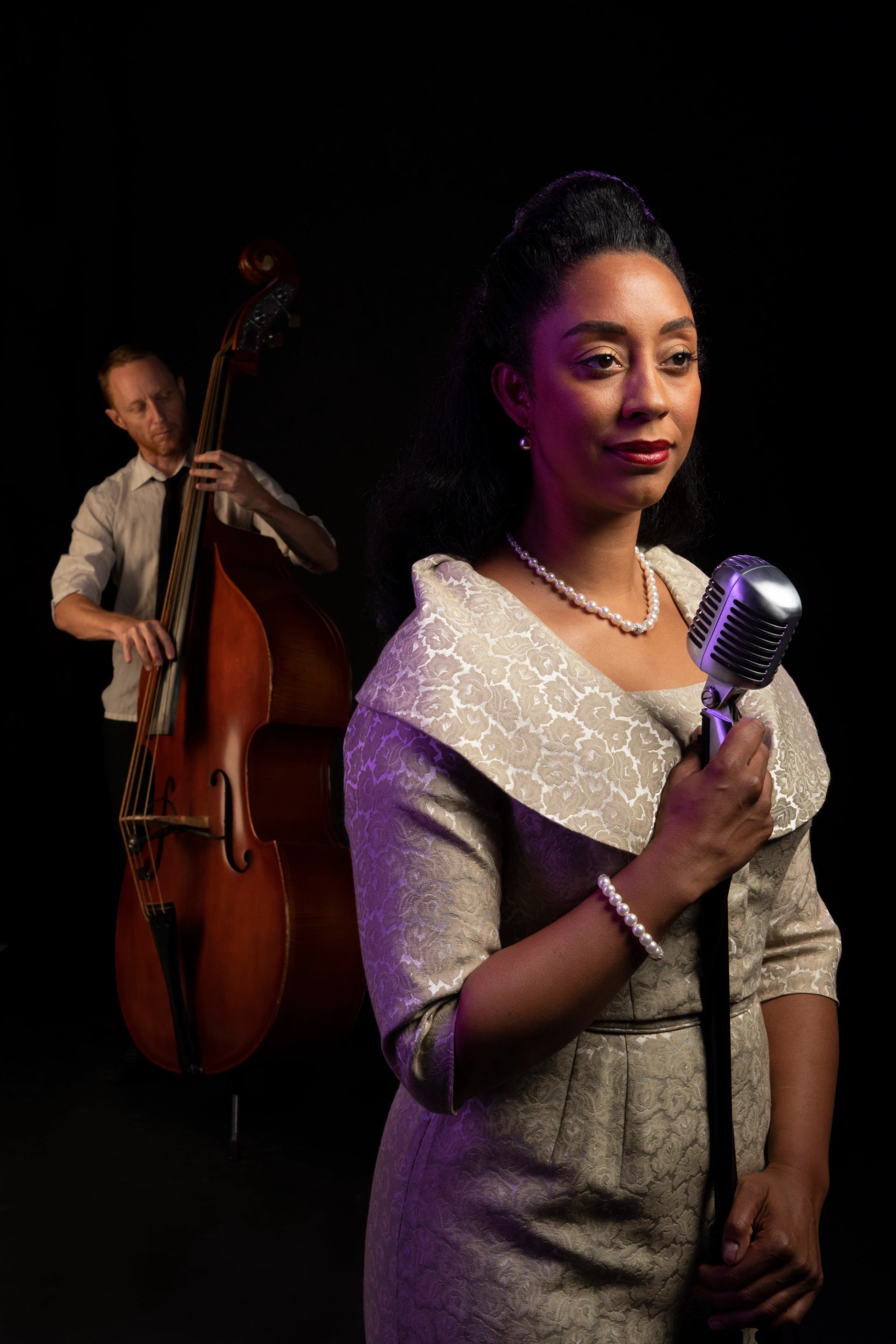 Jazz singer holding vintage microphone with bassist playing upright bass in dark studio.