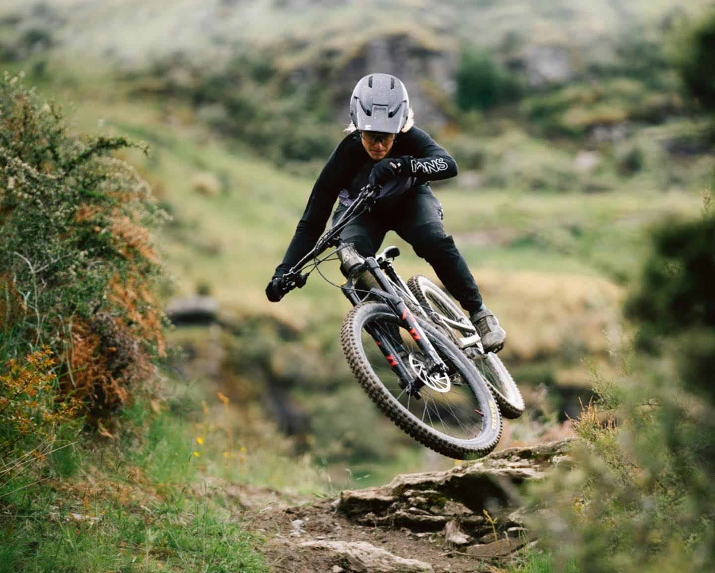 Mountain biker airborne on rugged trail surrounded by greenery and rocks.