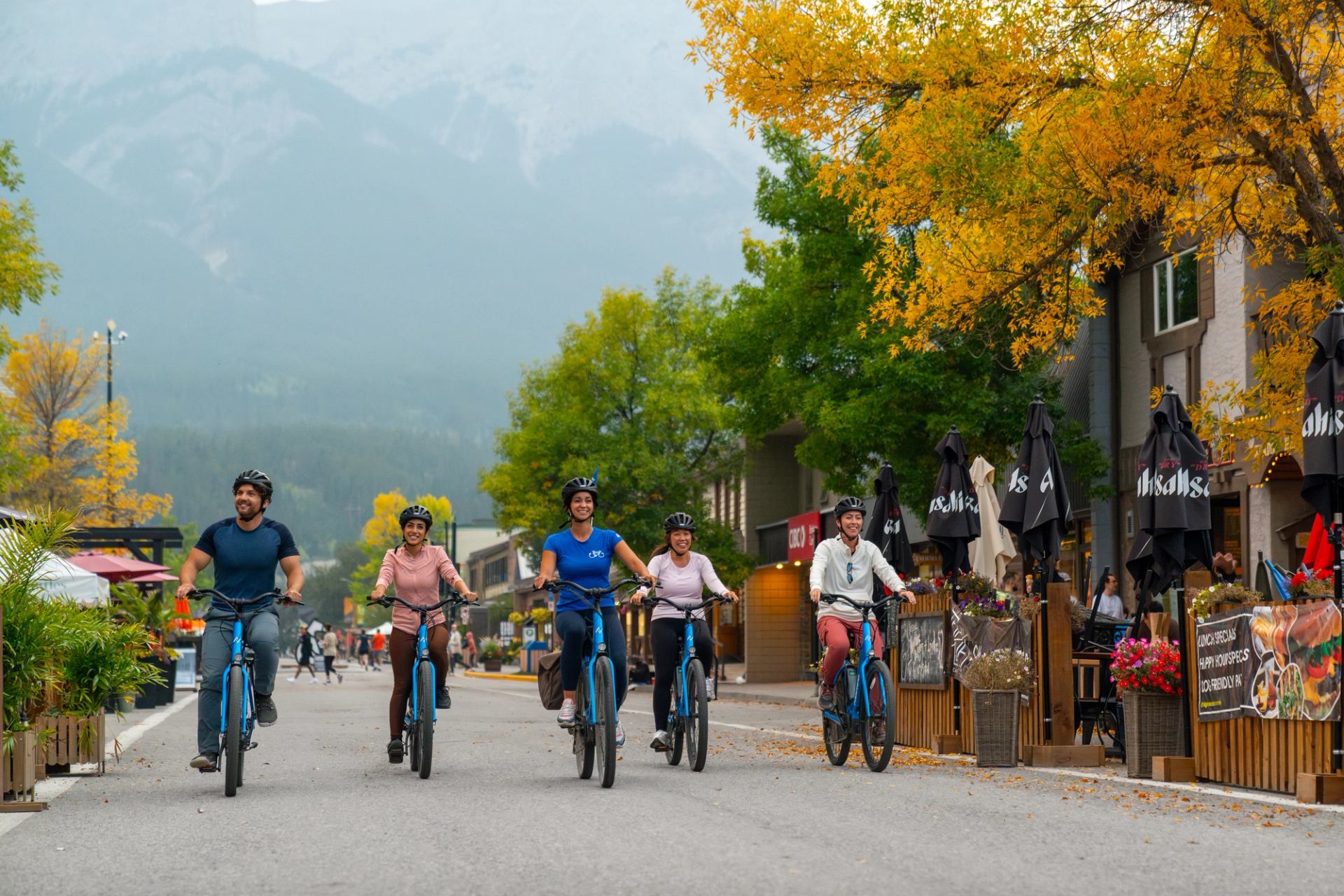 Five people ride blue bicycles down an autumn street lined with shops, with mountains in the background.