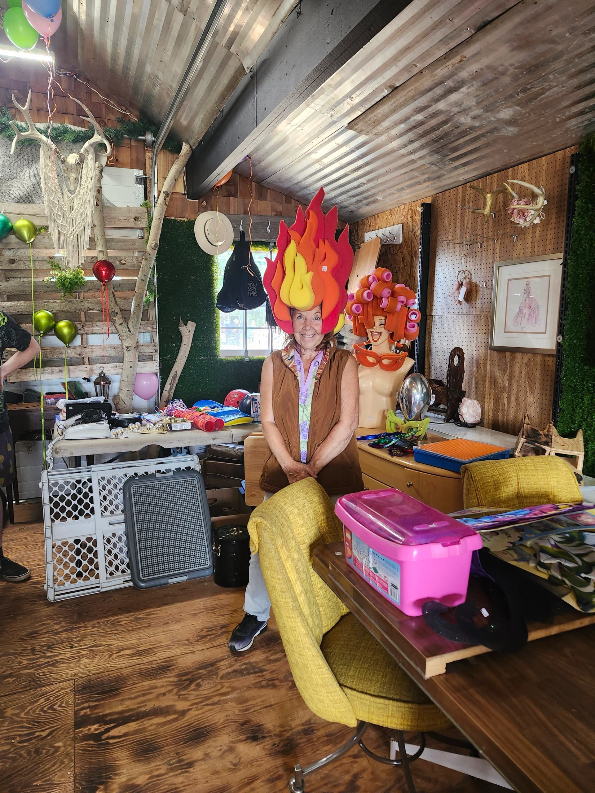 Person wearing large flame-shaped hat stands in cluttered rustic room with decorations.