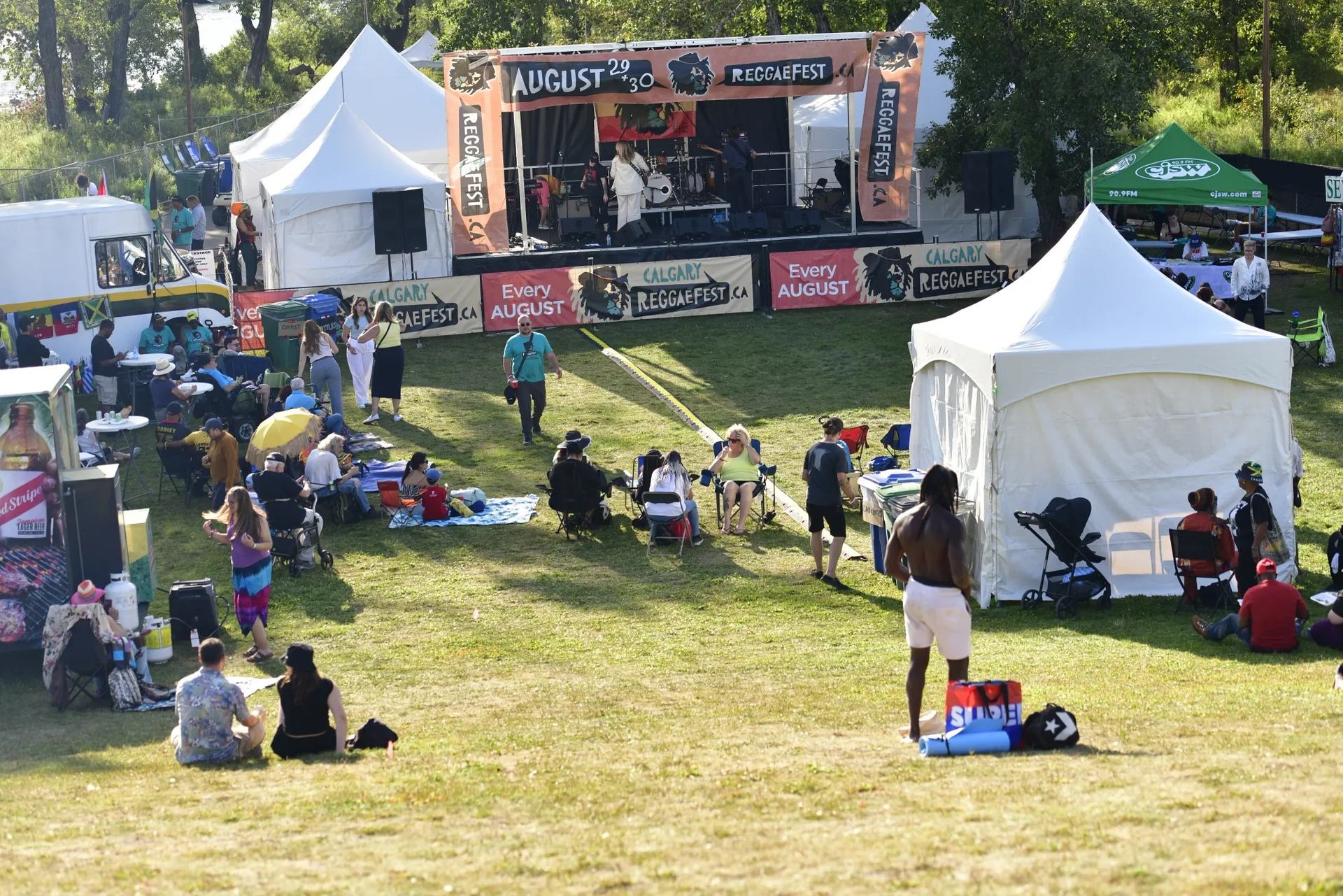 Outdoor reggae festival crowd sitting on grass facing stage with tents and banners.