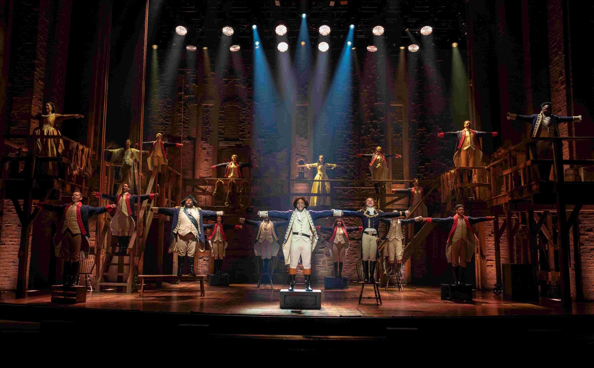 Stage ensemble in period military costumes extends arms under bright spotlights on wooden set