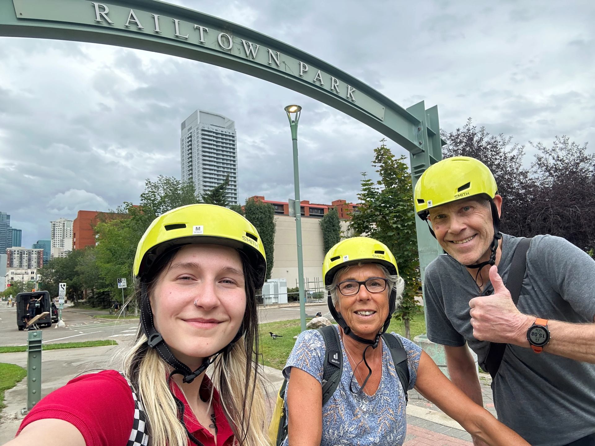 Three helmeted people take a selfie at Railtown Park with buildings and trees behind.