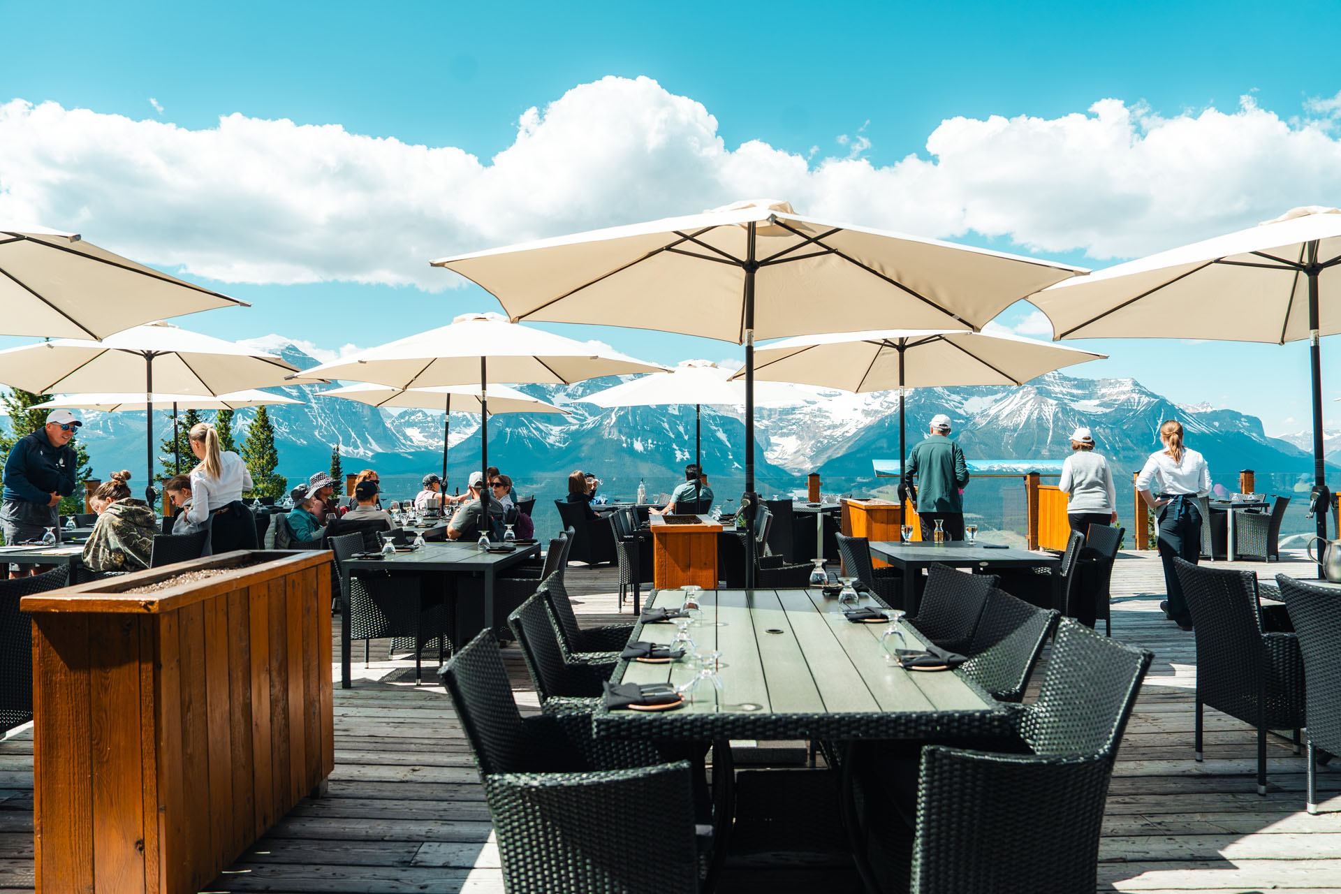 Outdoor restaurant patio with umbrellas overlooking snow-capped mountain range and blue sky.