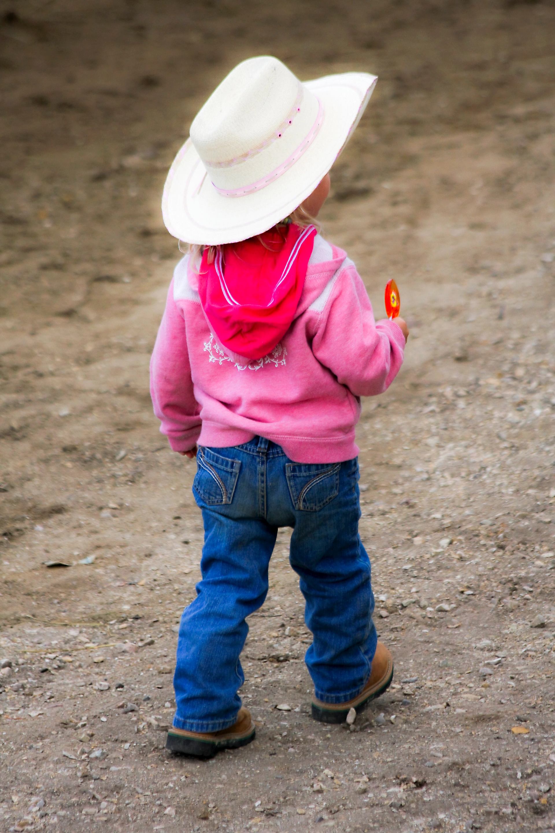 Small child in western clothing walking across a dirt rodeo ground.