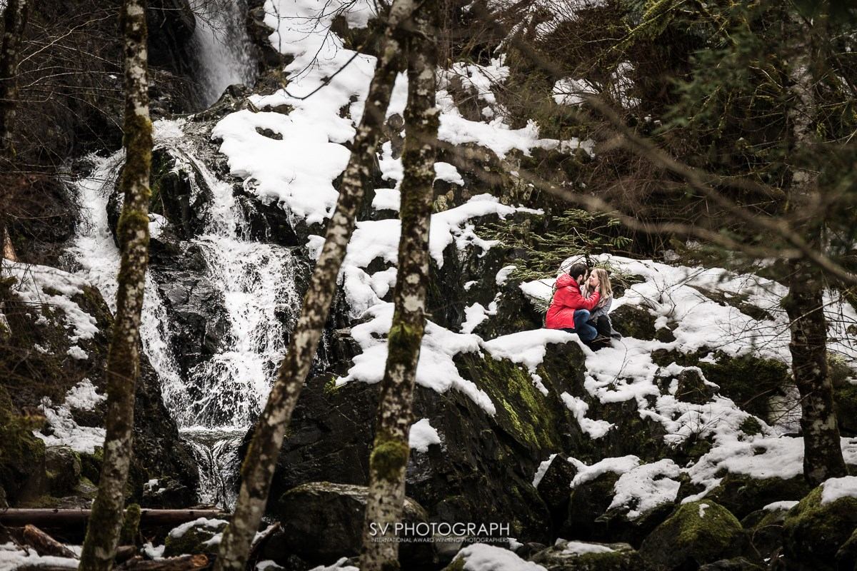 Person in red jacket sits on snowy rocks by a waterfall in a winter forest, framed by bare trees in serene peace.