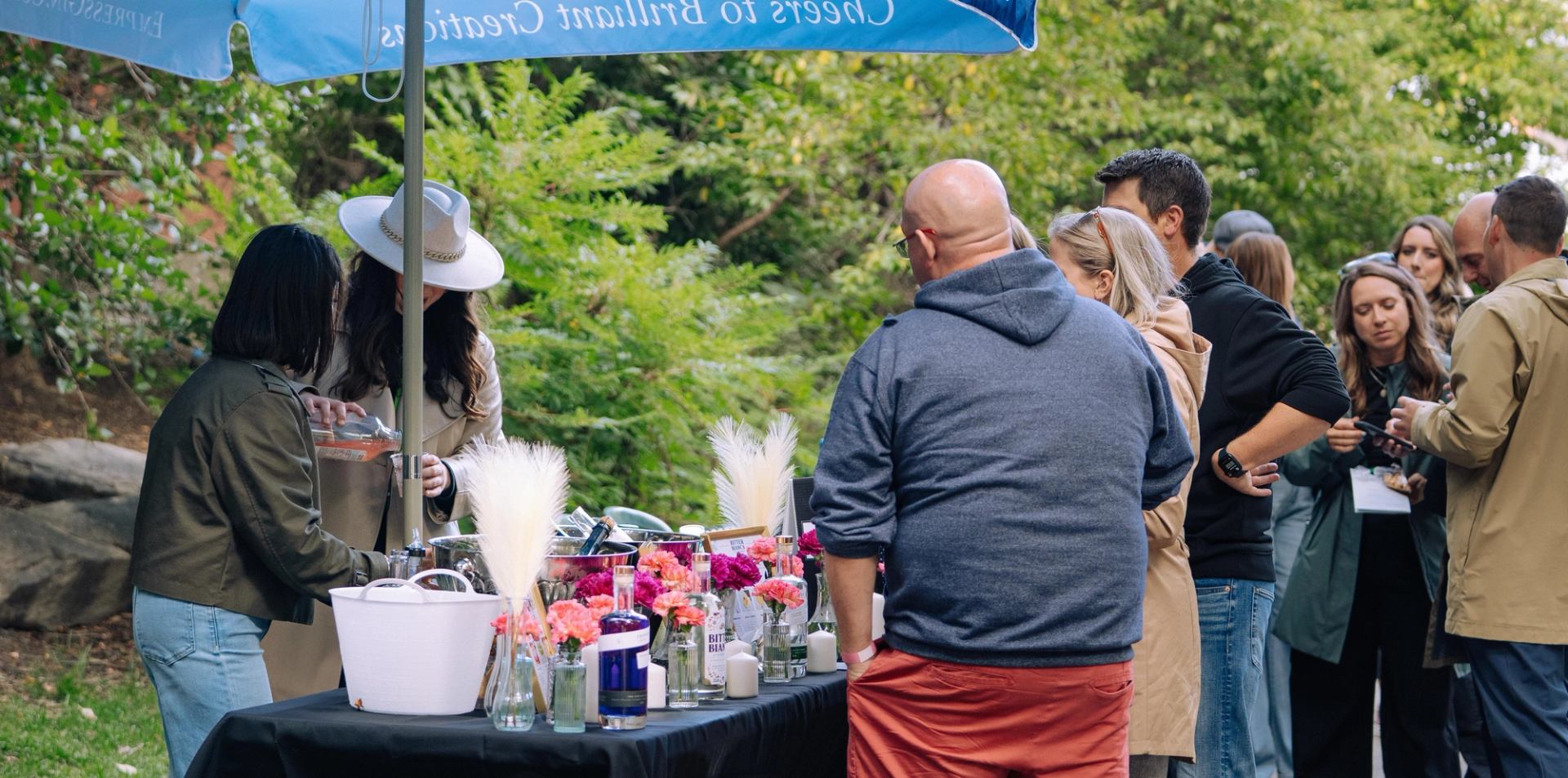 Outdoor gathering with people standing at a drink table under a branded umbrella.