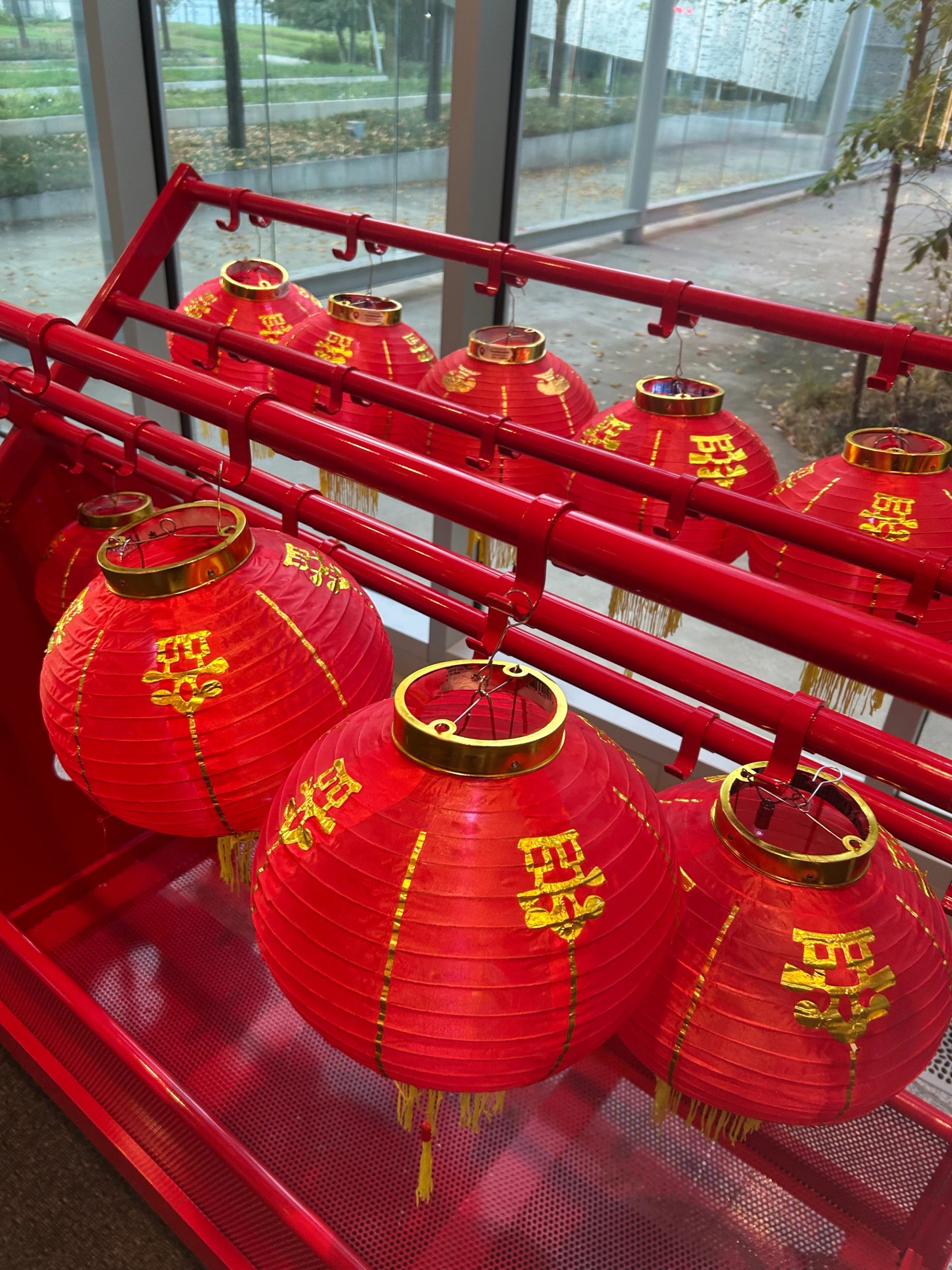Red Chinese lanterns with gold characters hang on red metal racks by windows.