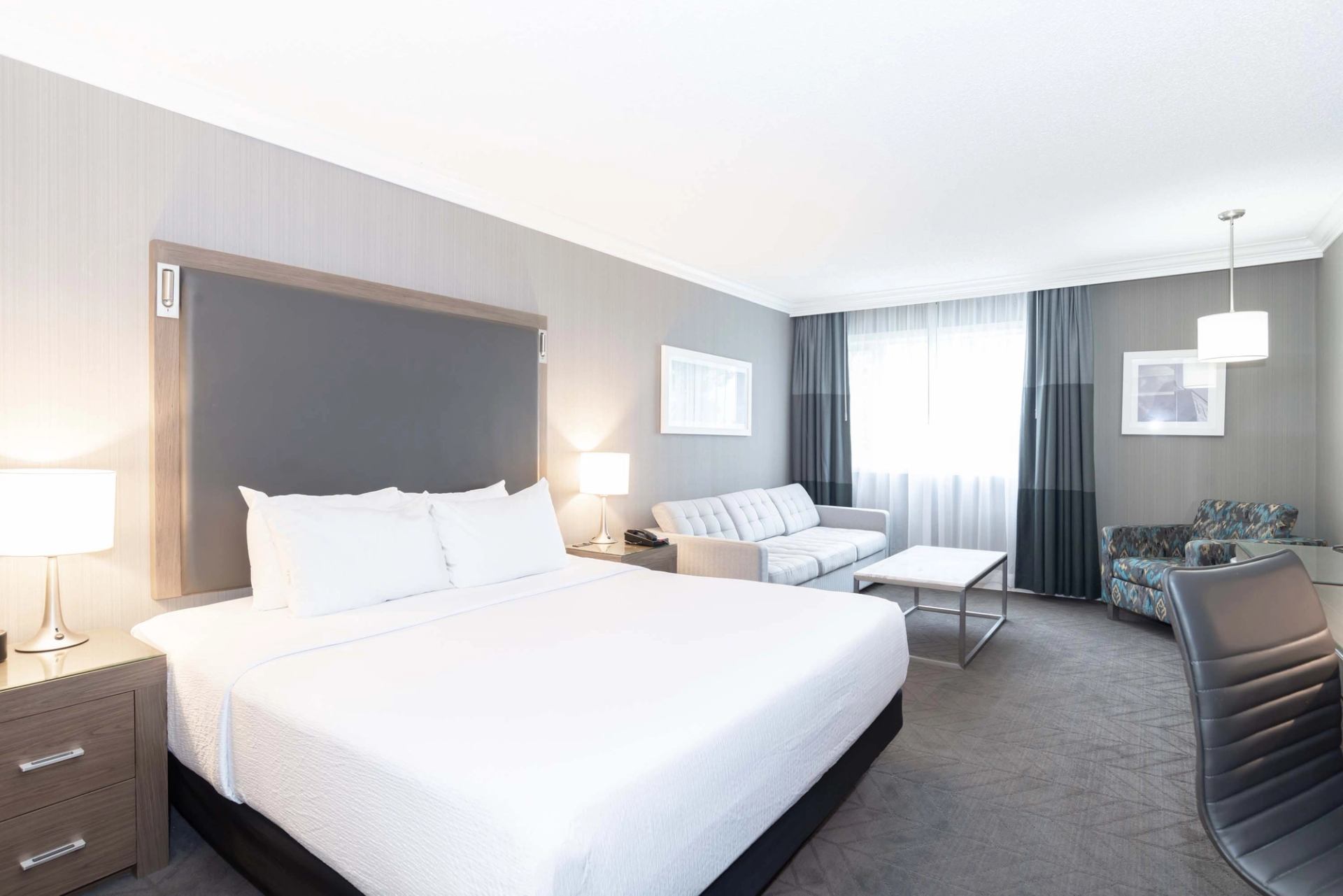 Hotel room with large bed, sofa, armchairs, desk chair and window with curtains.