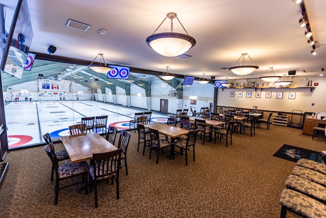A spacious lounge area with tables and chairs overlooking a multi-sheet curling rink through a glass partition.