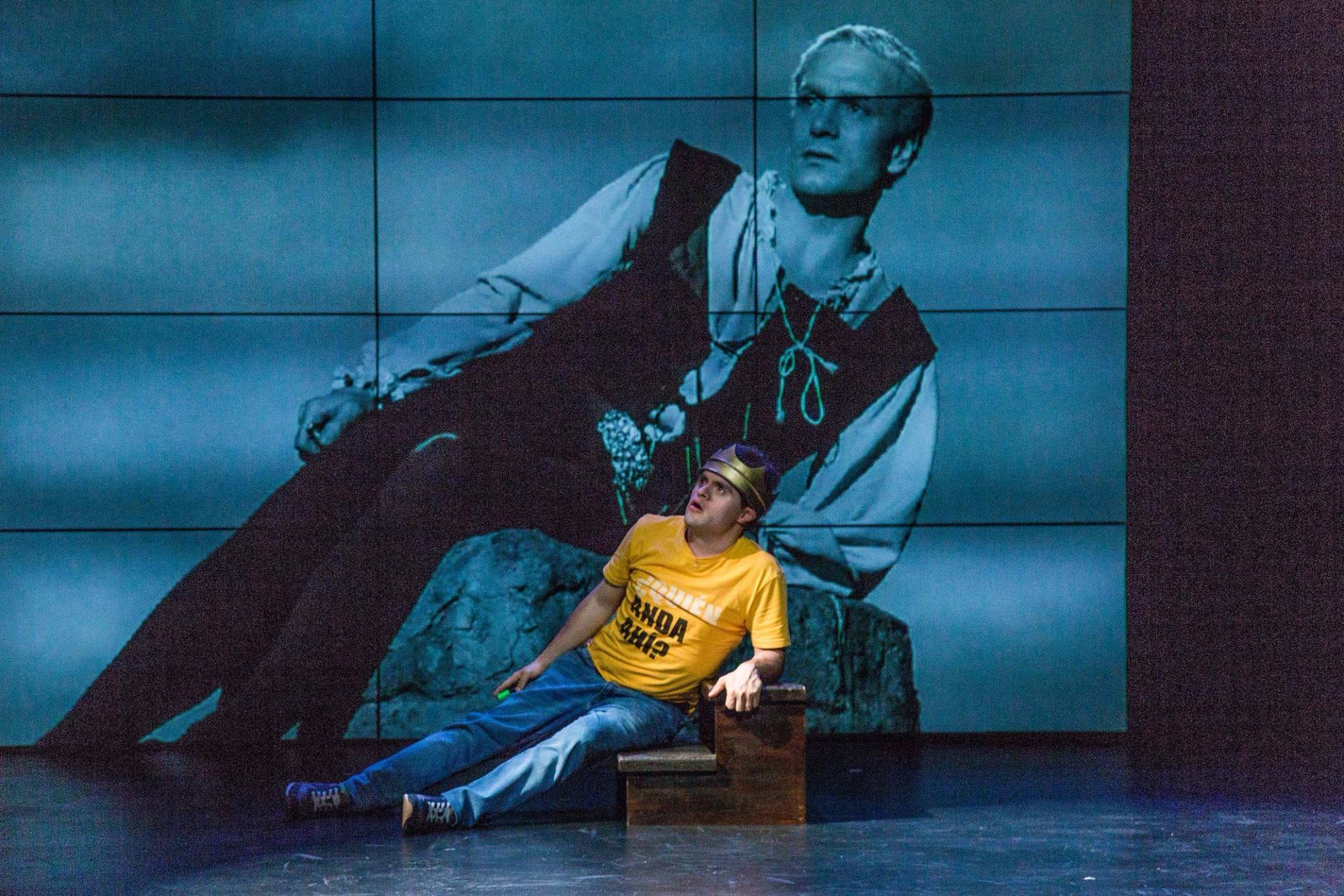 Performer in a yellow shirt sits before a large blue-tinted projected figure.