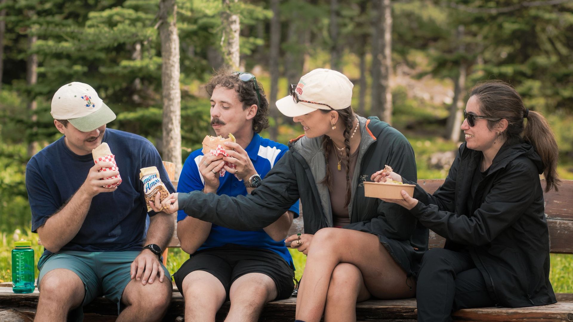 Group of people sitting on a bench outdoors, eating sandwiches and snacks together.