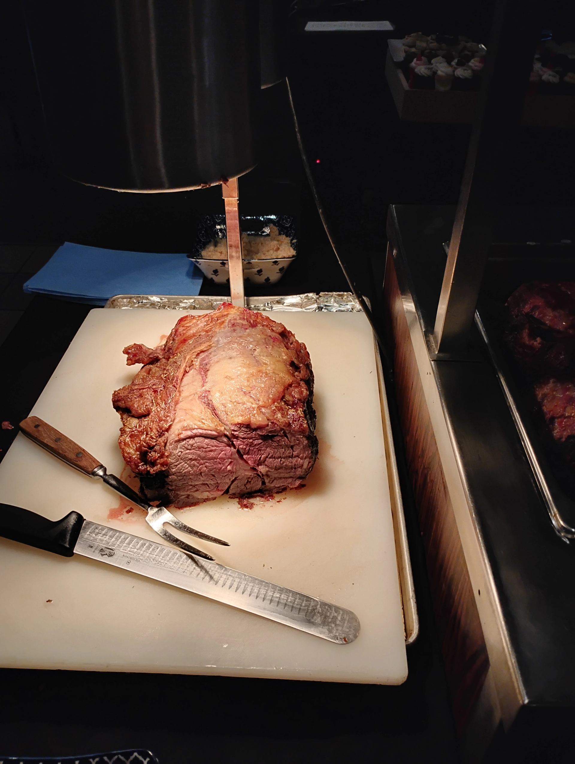 A large, roasted cut of meat on a white carving board with a knife and fork, illuminated by a heat lamp.