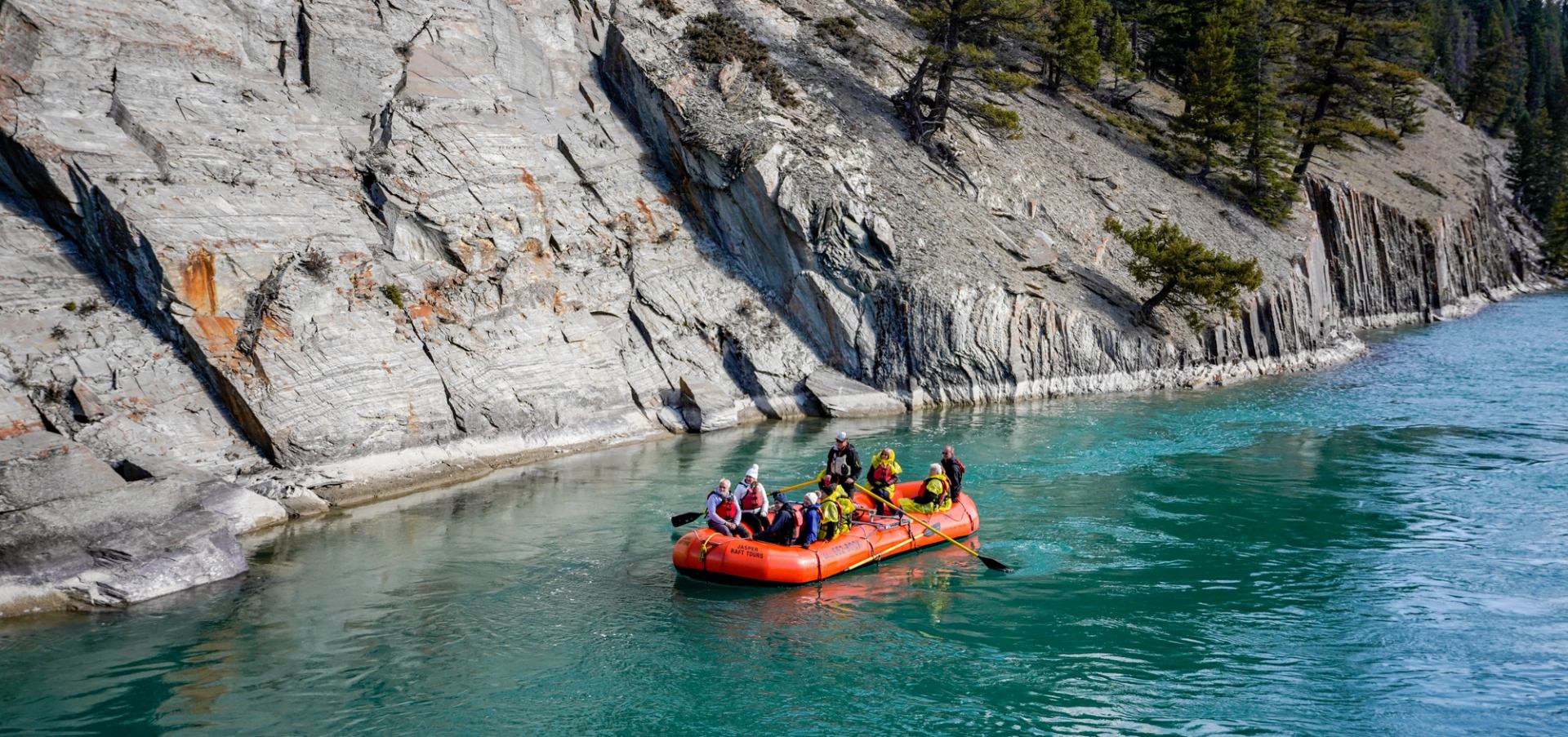 A red raft floats beside steep rocky canyon walls on clear blue-green water.