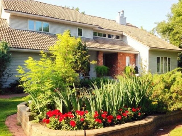 Large two-story house with a tiled roof, white siding, and a brick entryway surrounded by lush greenery and vibrant red flowers.