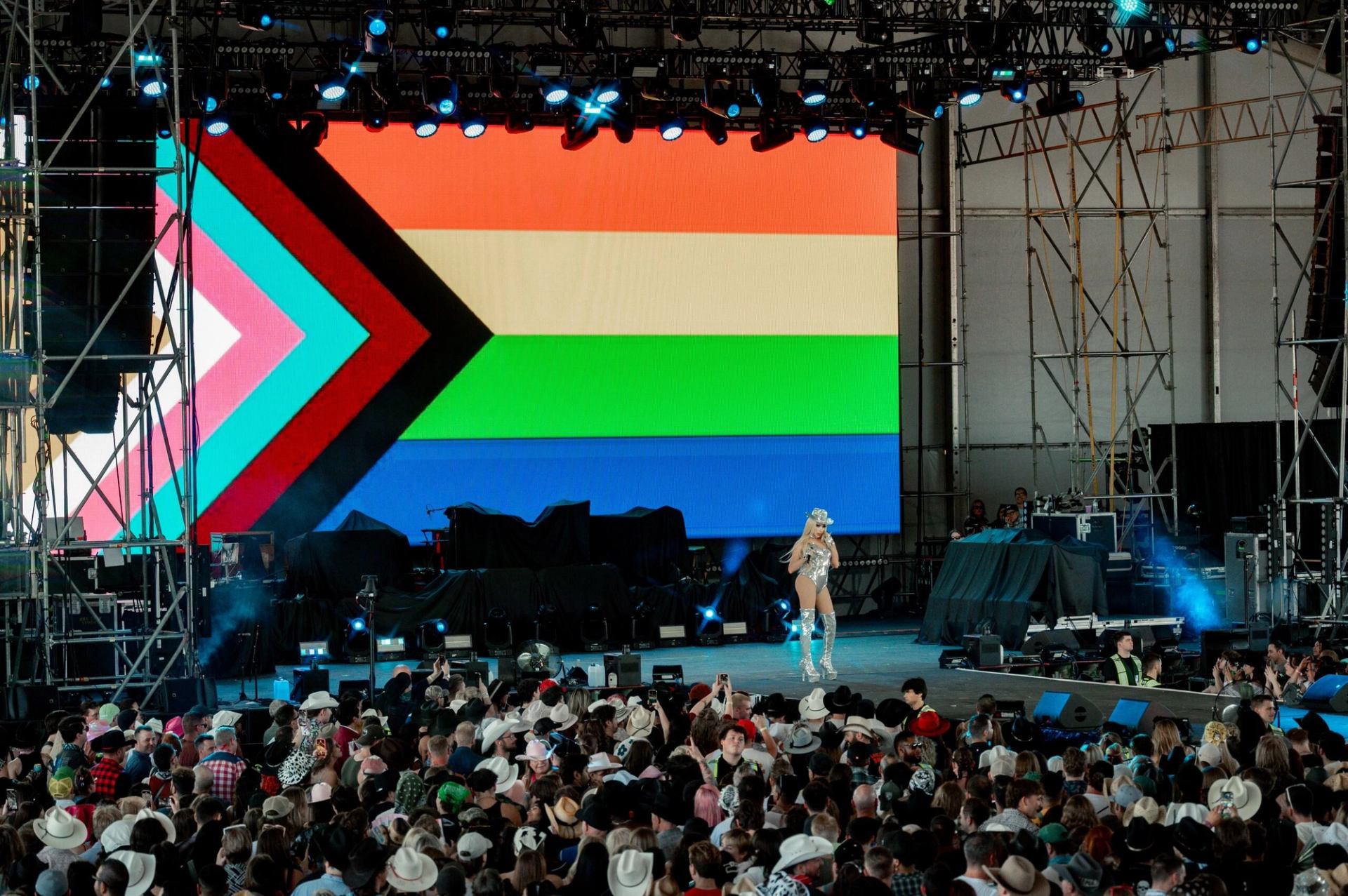 Performer in silver outfit on stage before large Progress Pride flag screen and crowd.