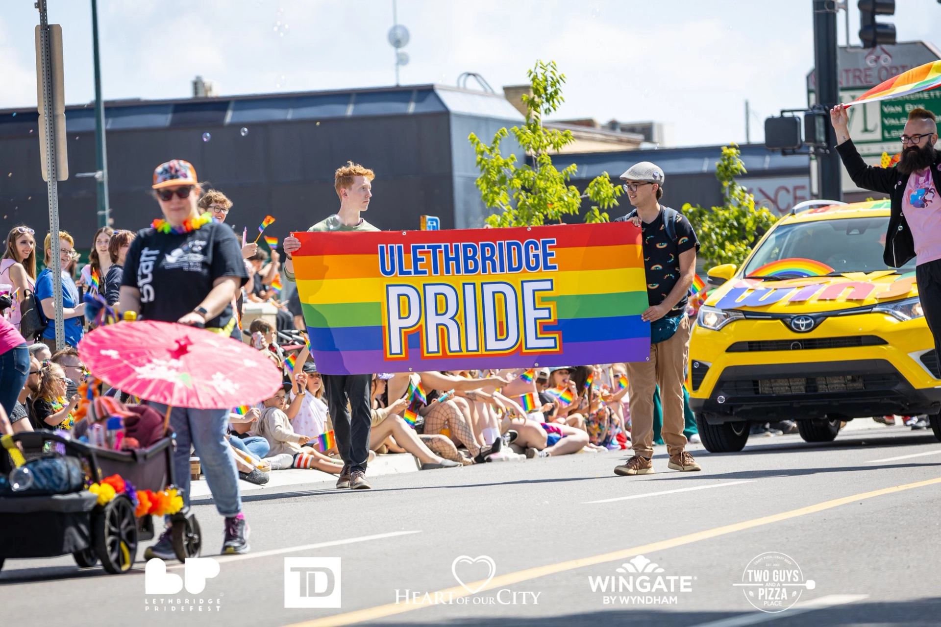 People carrying a Lethbridge Pride banner during a busy Pride parade with crowds watching.
