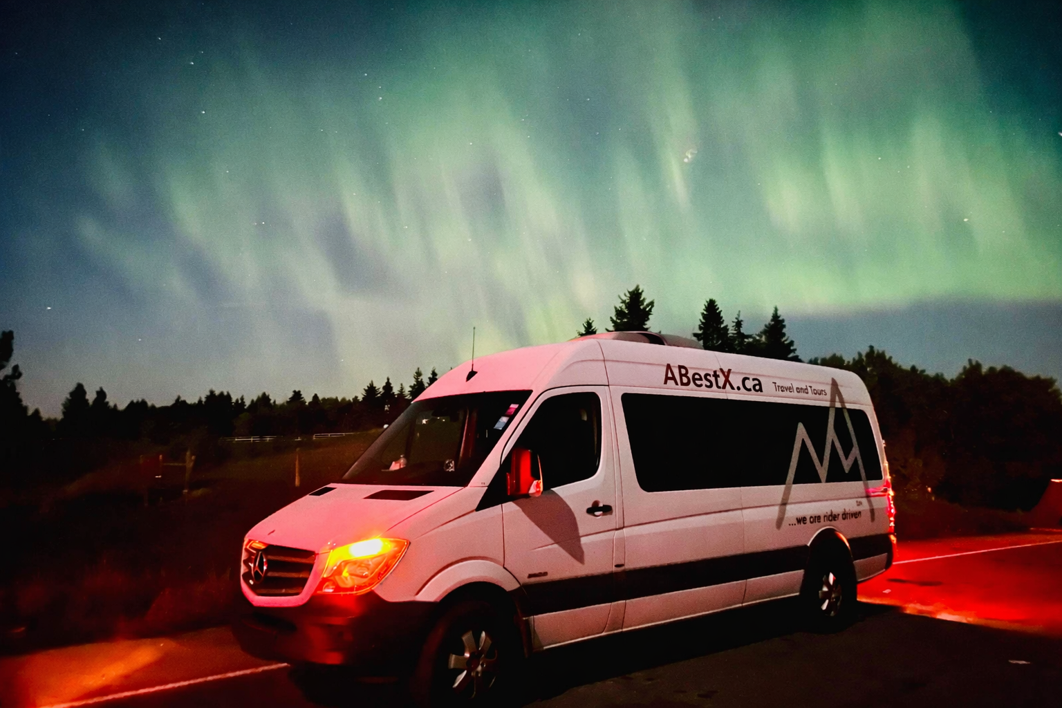 Tour van with ABestX.ca branding parked under green aurora beside dark treeline.