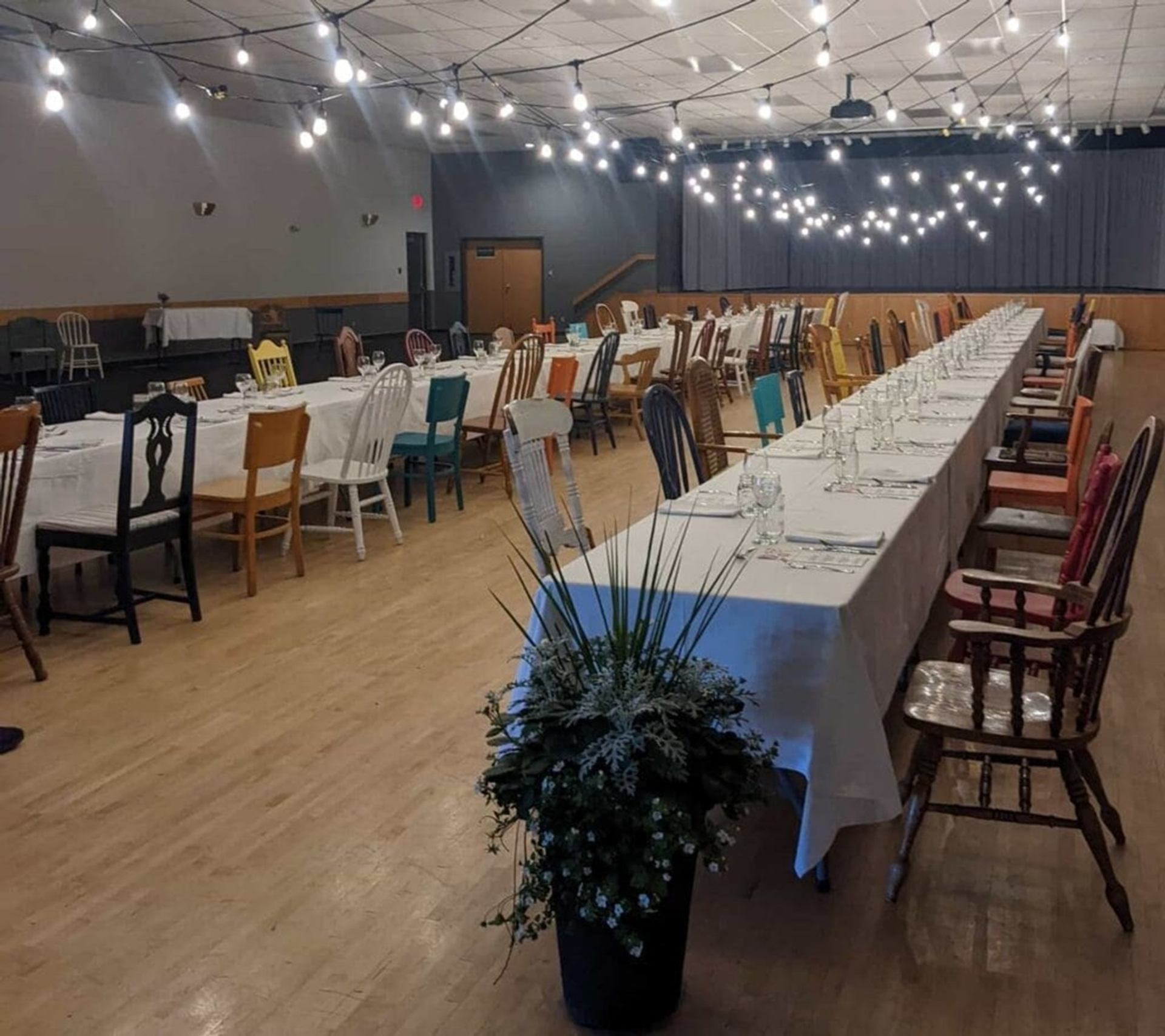 Banquet hall with long tables, mismatched wooden chairs and string lights overhead.