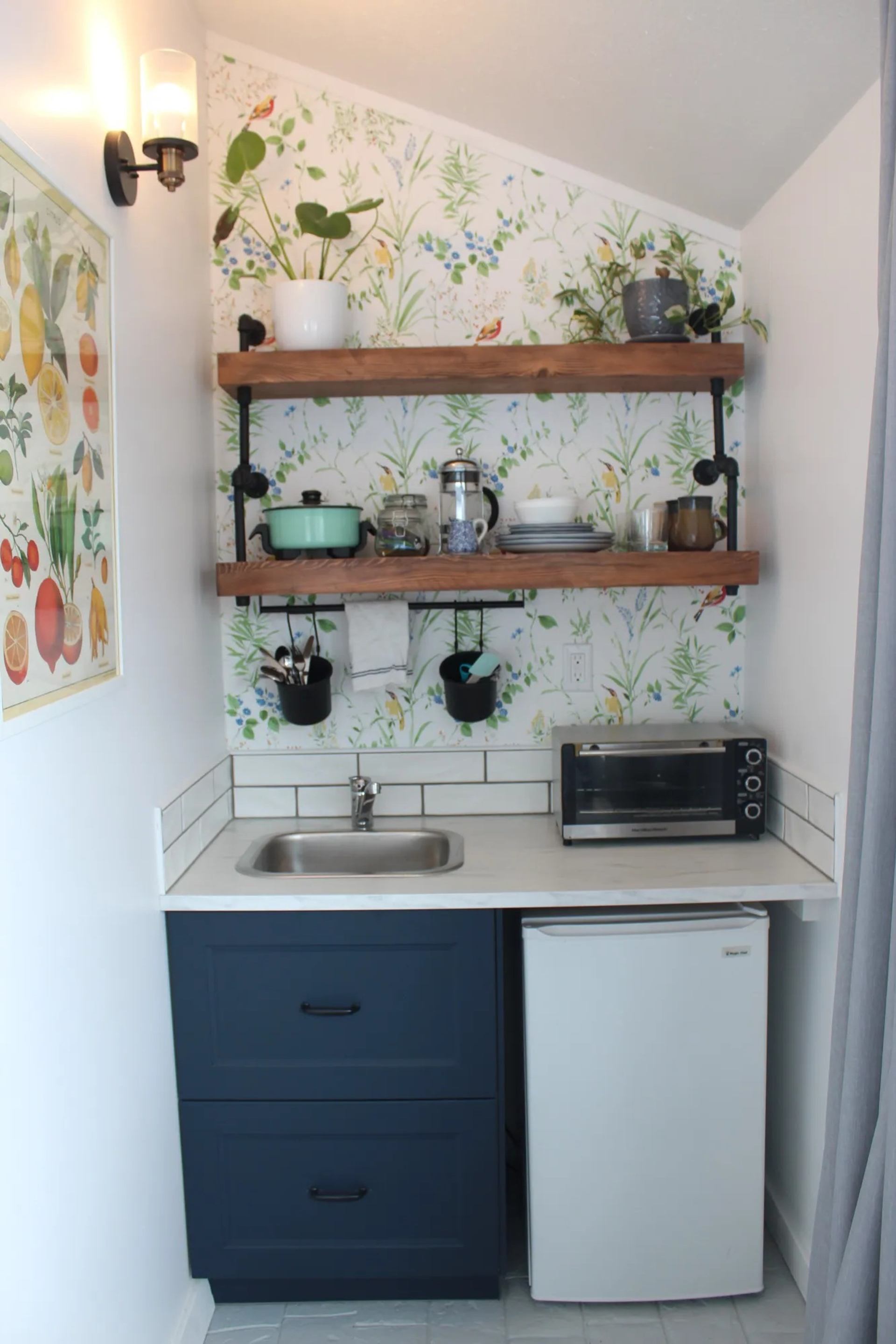 Compact kitchenette with sink, mini fridge, open shelves, toaster oven and plants.
