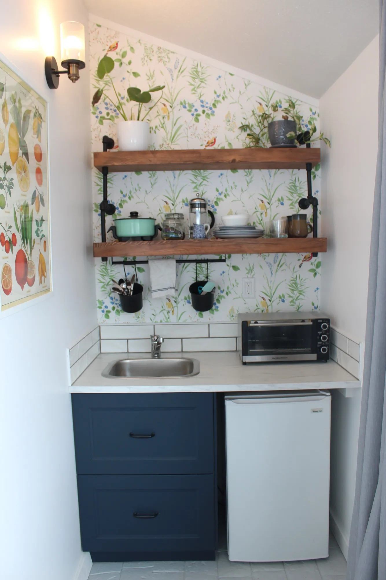 Compact kitchenette with sink, mini fridge, open shelves, toaster oven and plants.