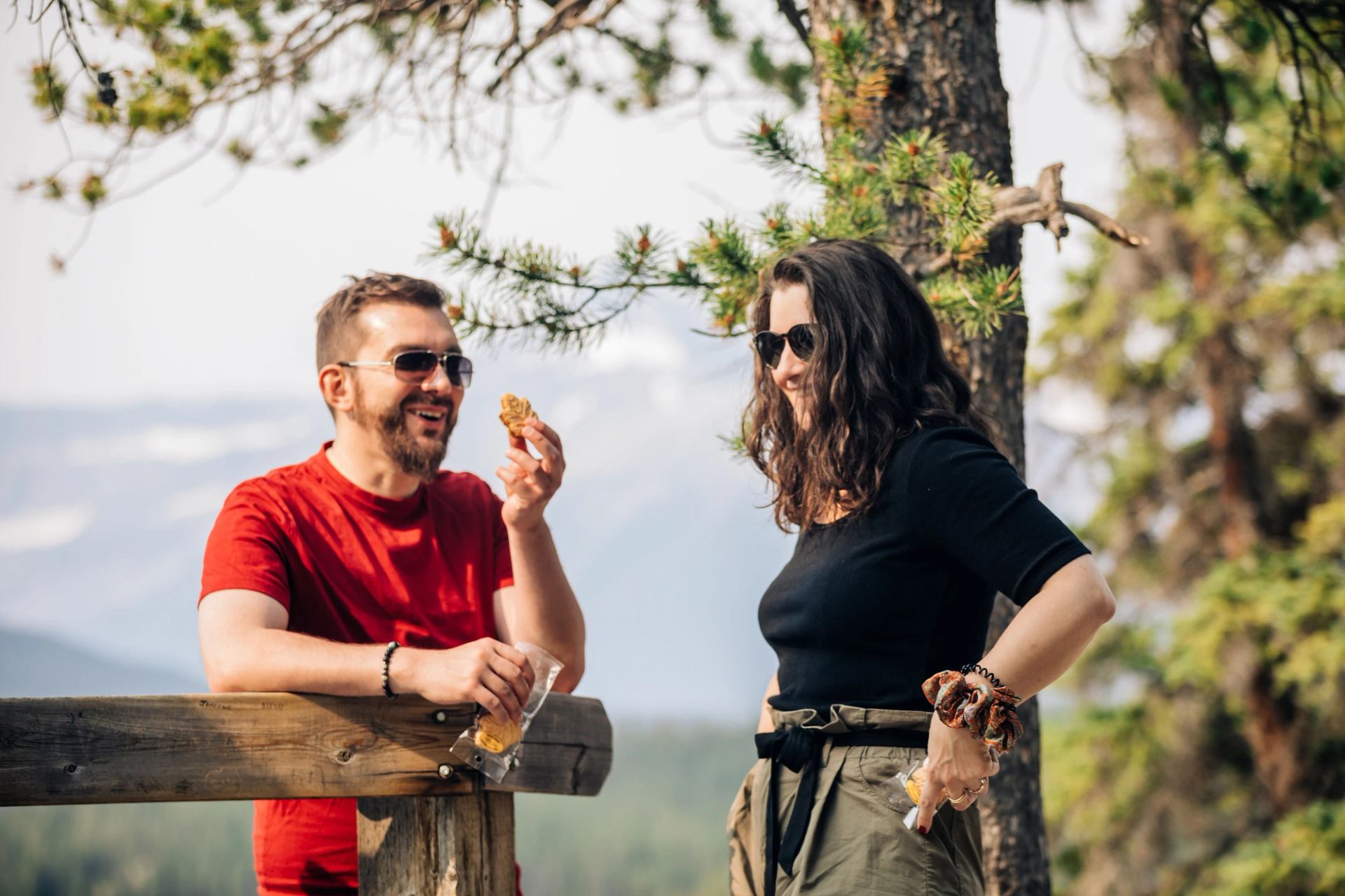 Two people wearing sunglasses share snacks by a wooden railing in a forested area.
