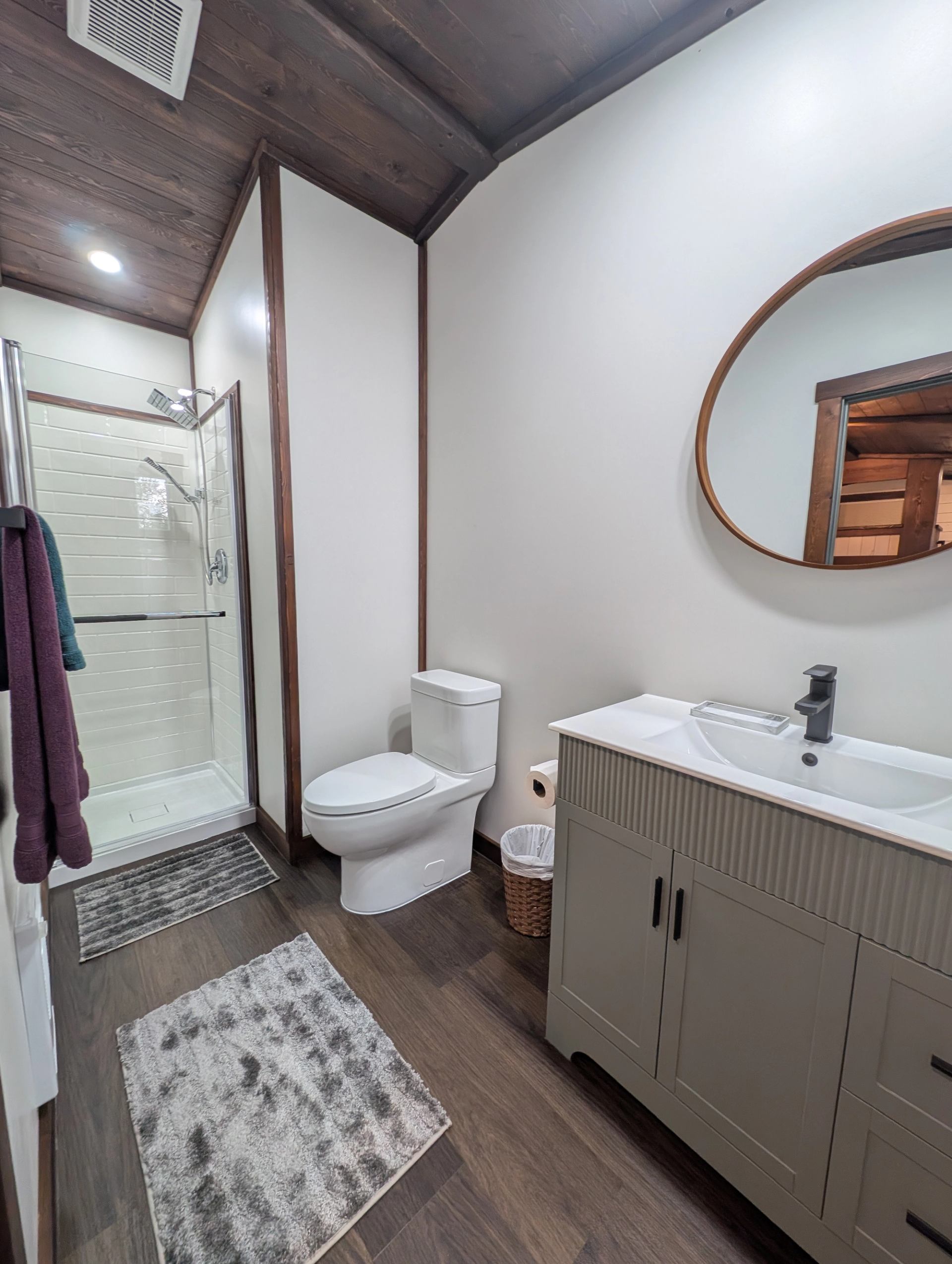 Bathroom with glass shower, toilet, grey vanity, round mirror and wood-panelled ceiling.