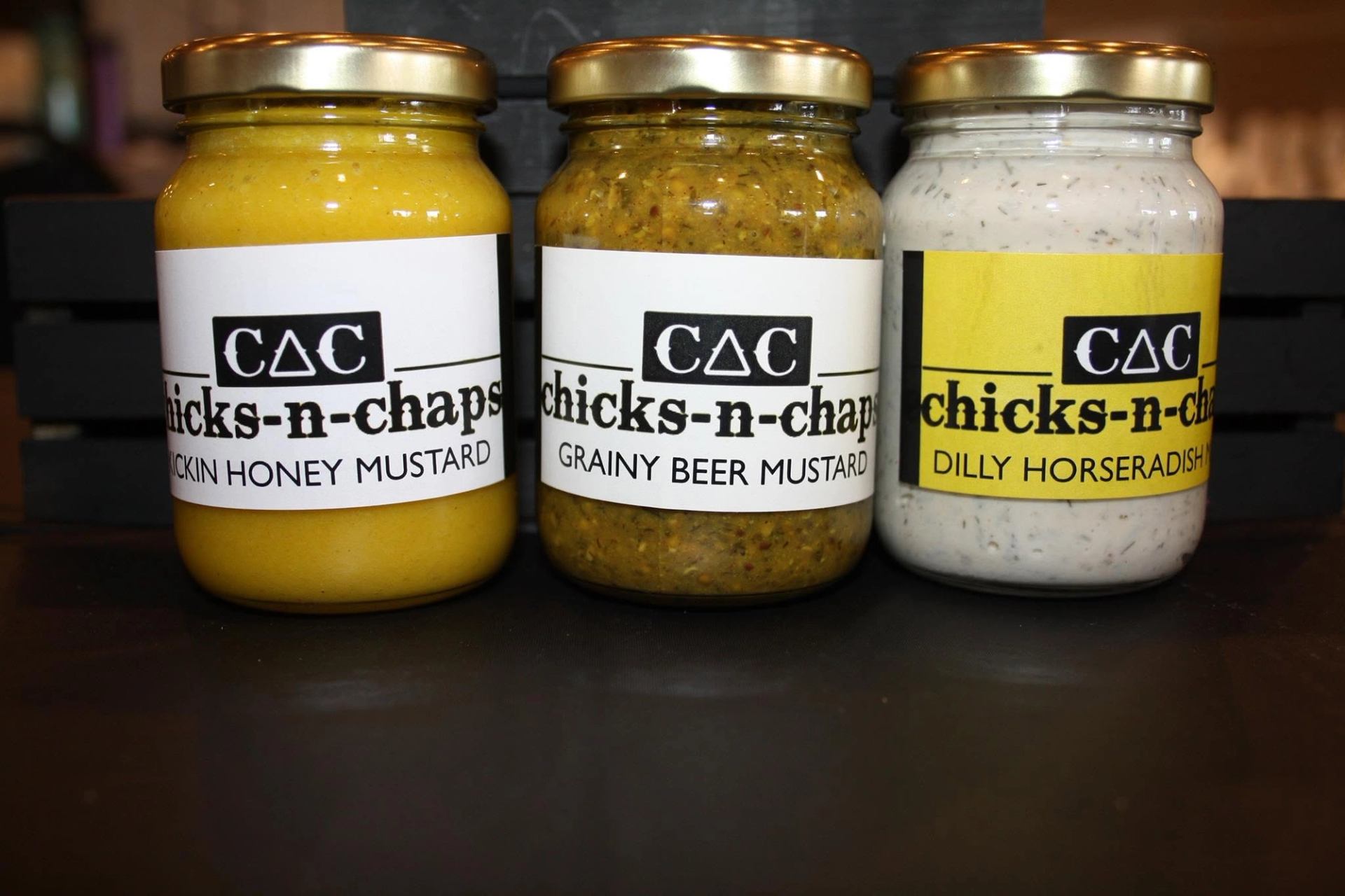 Three glass jars of specialty sauces and condiments are displayed on a wooden shelf.