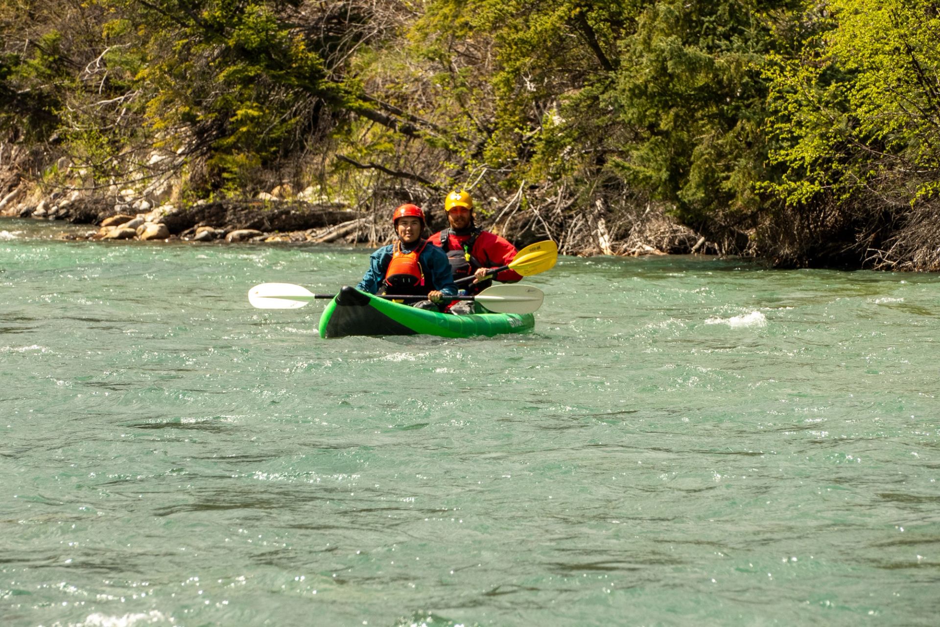 Two people paddle a green kayak down a fast-moving river beside forested banks.