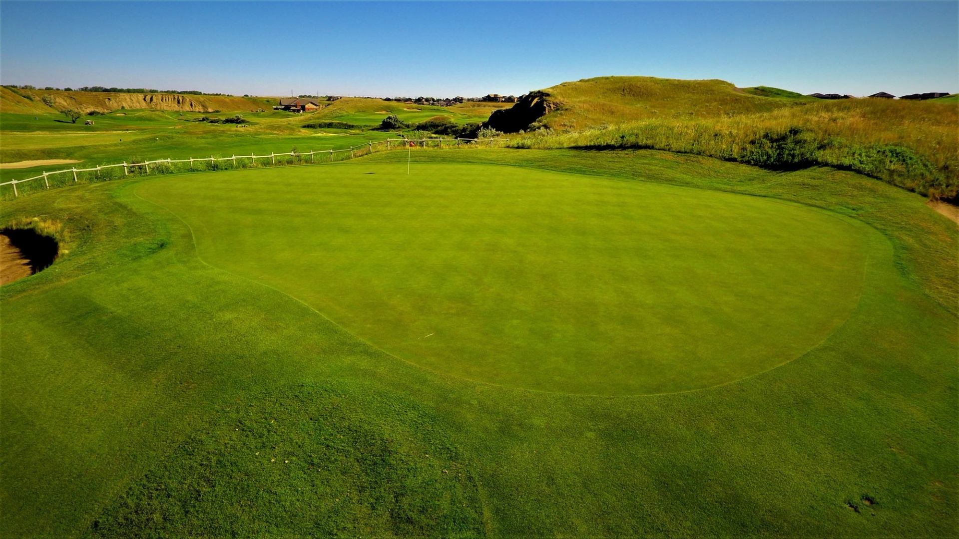 Golf green with flagstick beside rolling grass hills under clear blue sky.