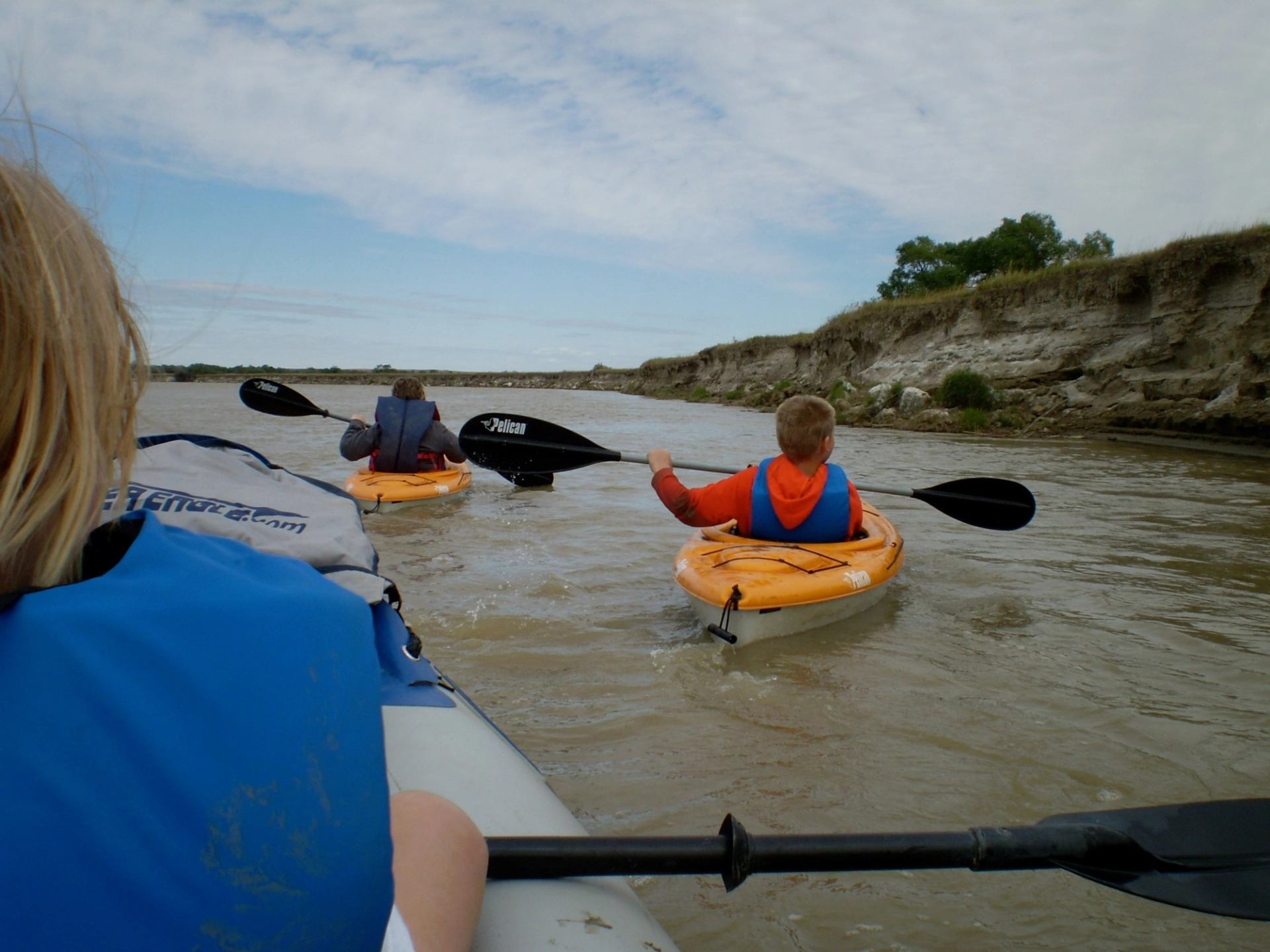 Kayakers paddle down a calm river near eroded cliffs under a partly cloudy sky.