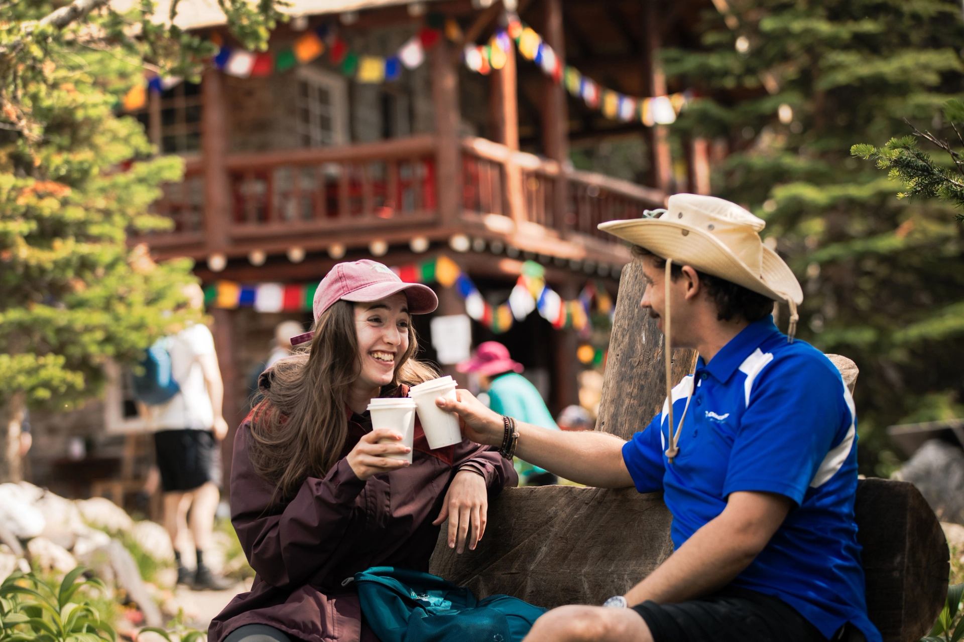 Two people sit outdoors with coffee cups near a rustic lodge and colourful flags.