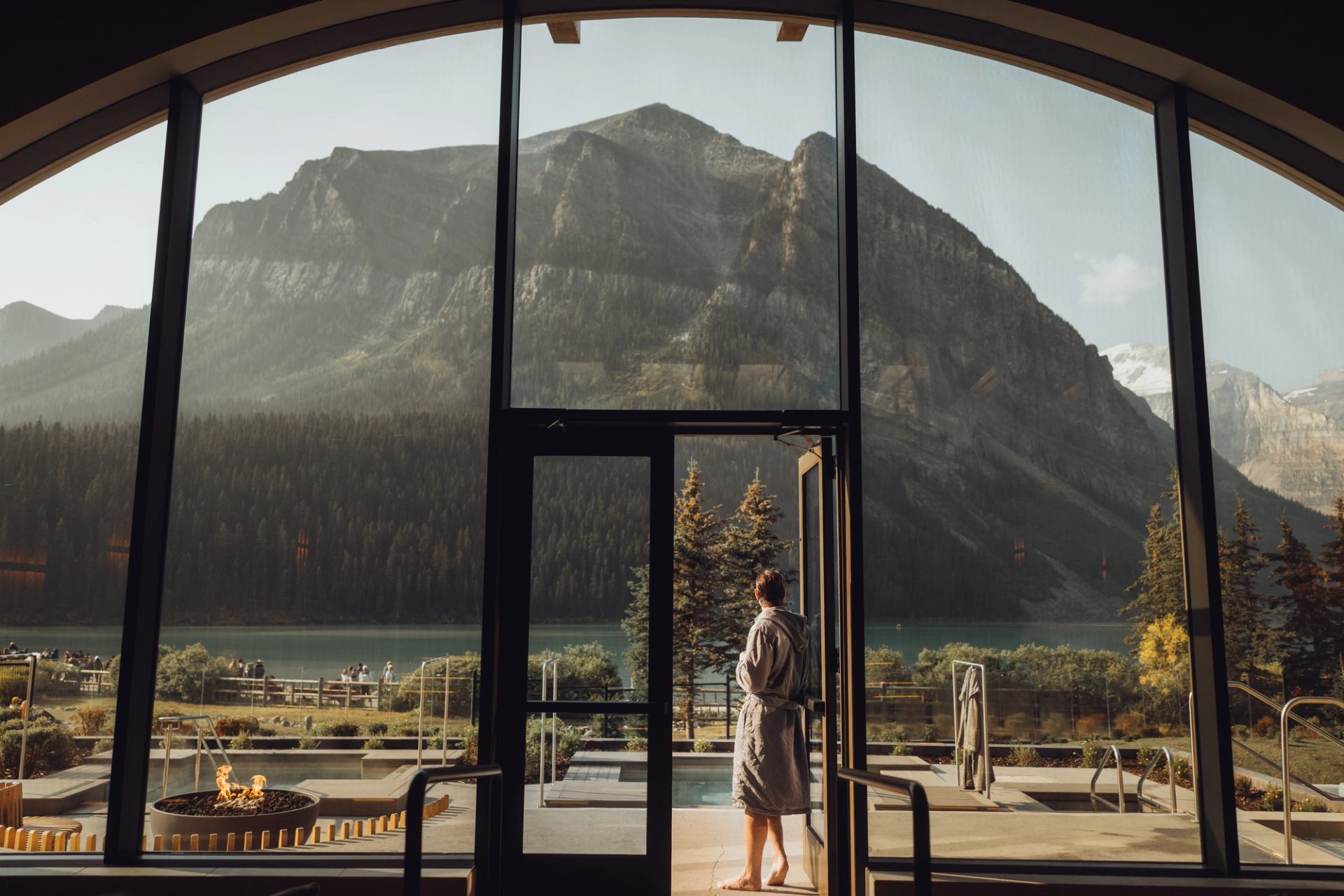A robed person looks out the glass doors of a mountain spa retreat.