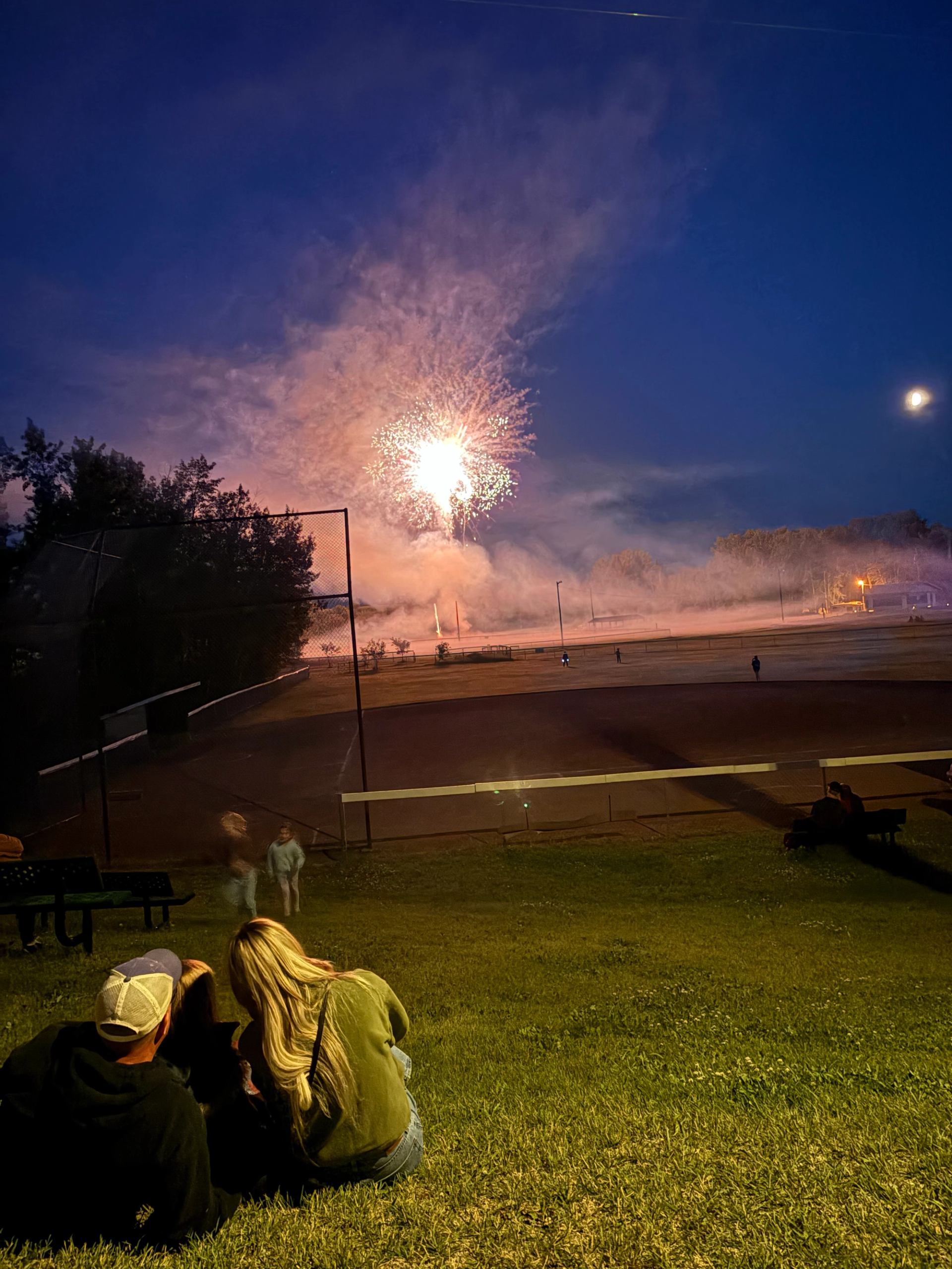 People sitting on grassy hill watching fireworks over sports field under dark sky.