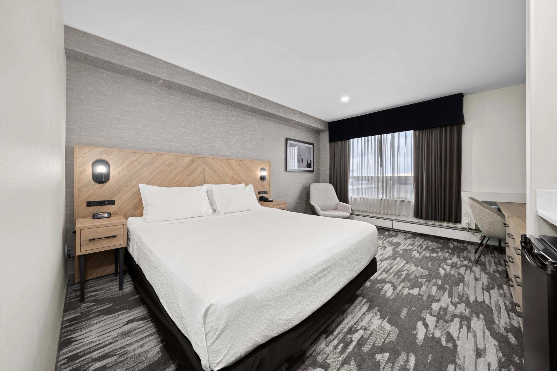 Hotel room with king bed, armchair, desk, window with curtains and carpeted floor.