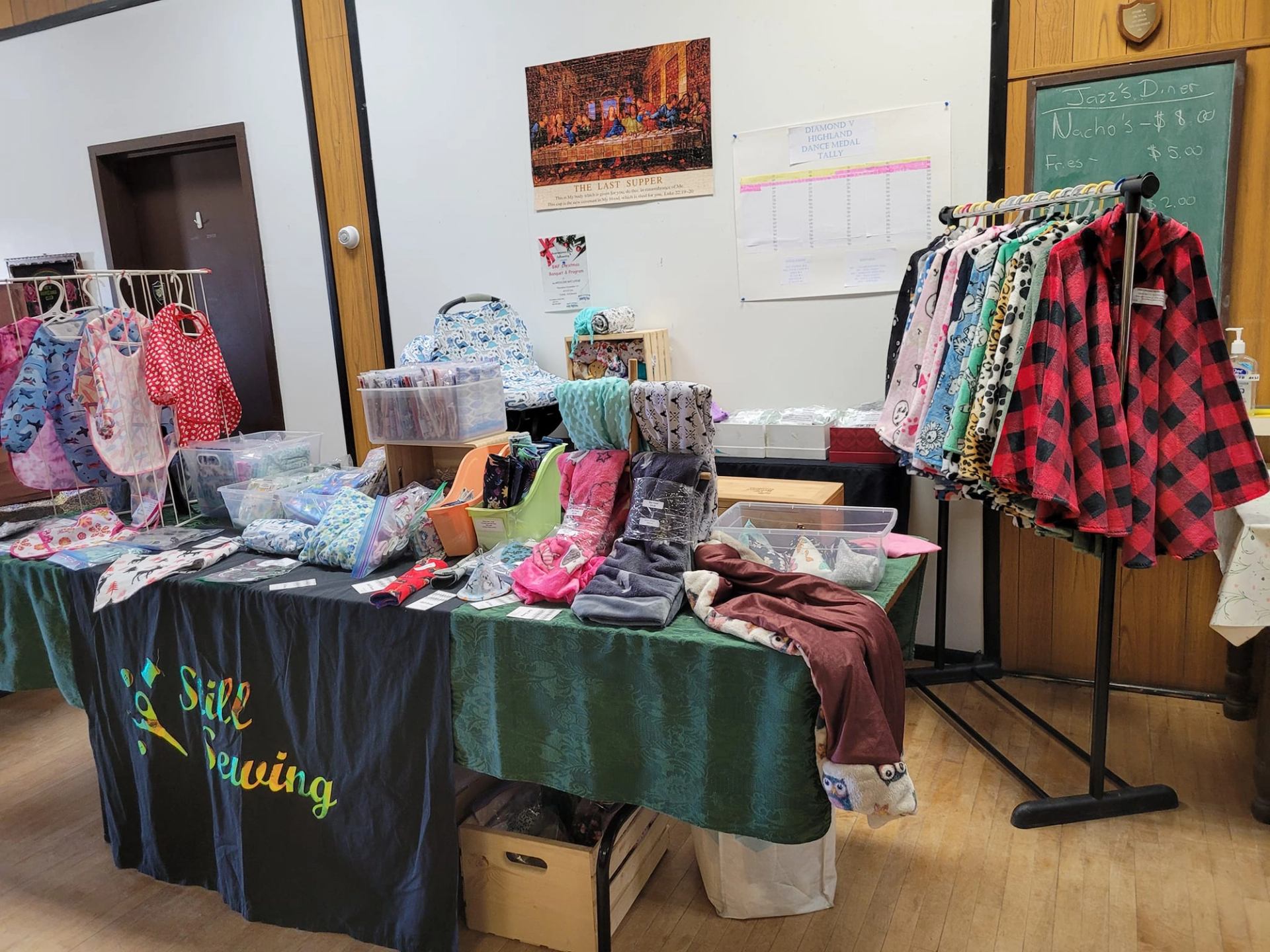 A craft fair booth displaying handmade baby clothing, bibs, patterned fabric items, and a rack of plaid shirts, with a "Still Sewing" banner.