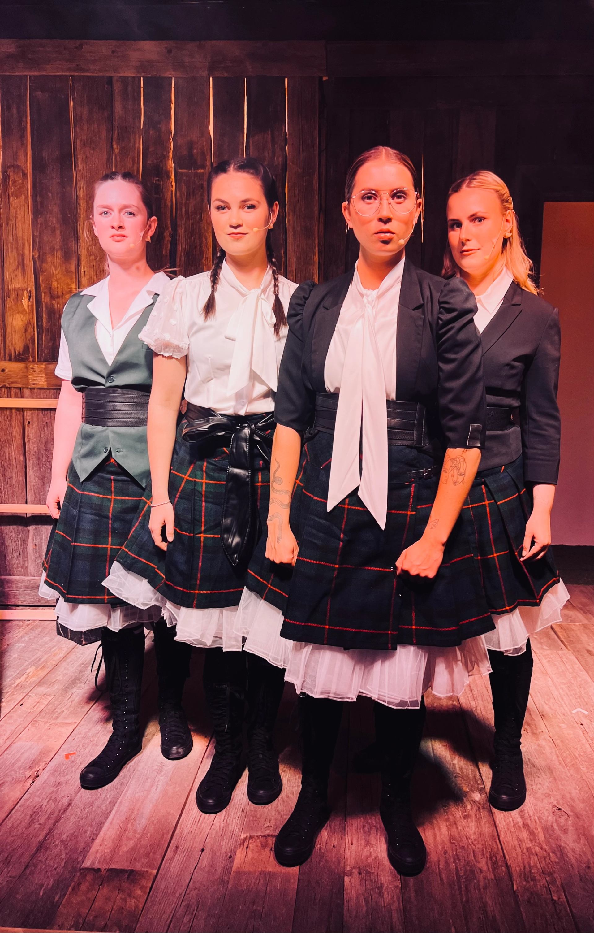 Four performers in plaid skirts pose on a warmly lit wooden stage.